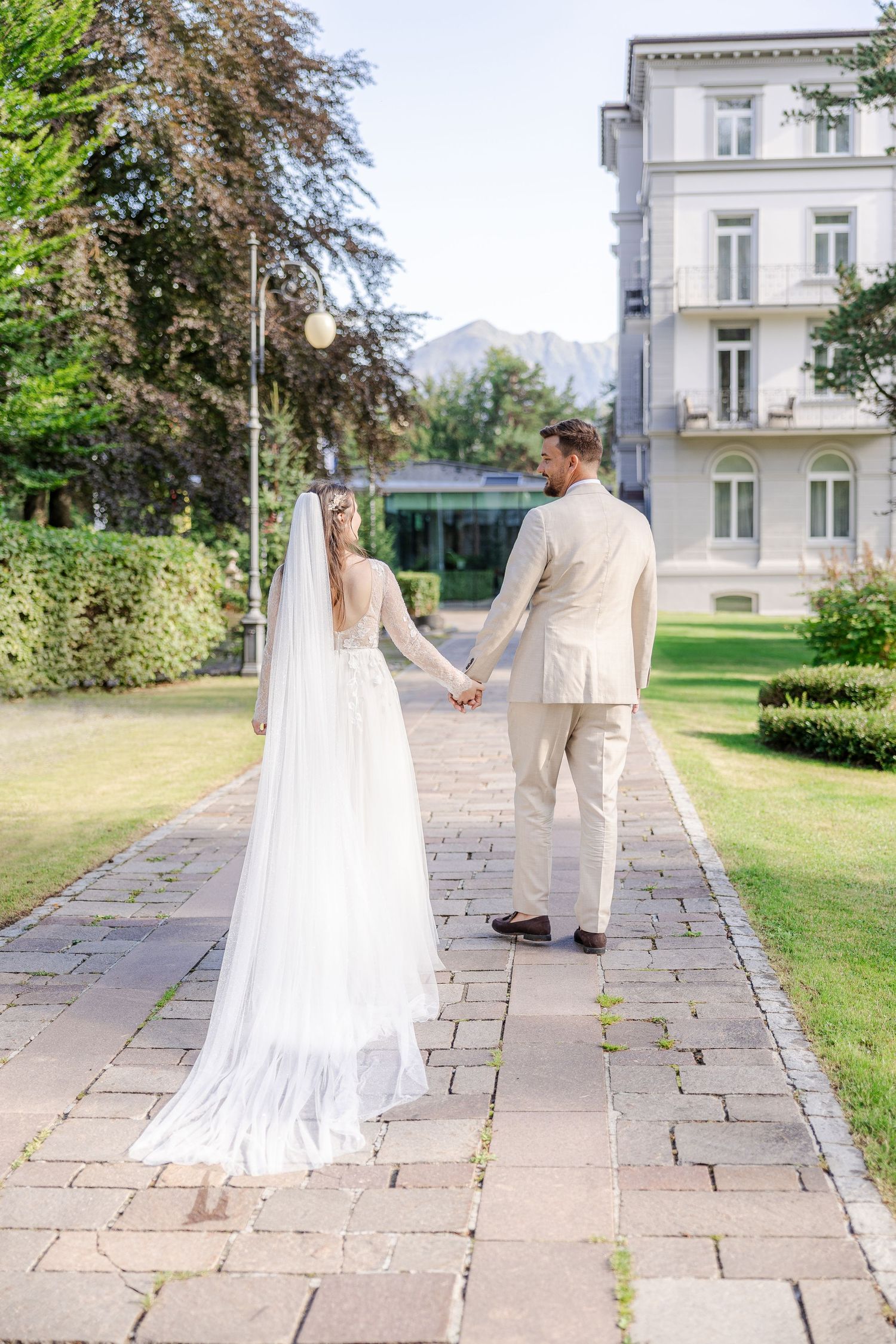 A couple in wedding attire walks along a stone path outside an elegant white building on their special day.