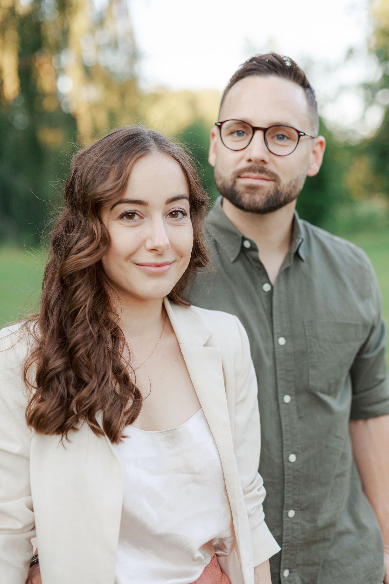 A couple poses outdoors in a park setting with natural lighting and greenery in the background.