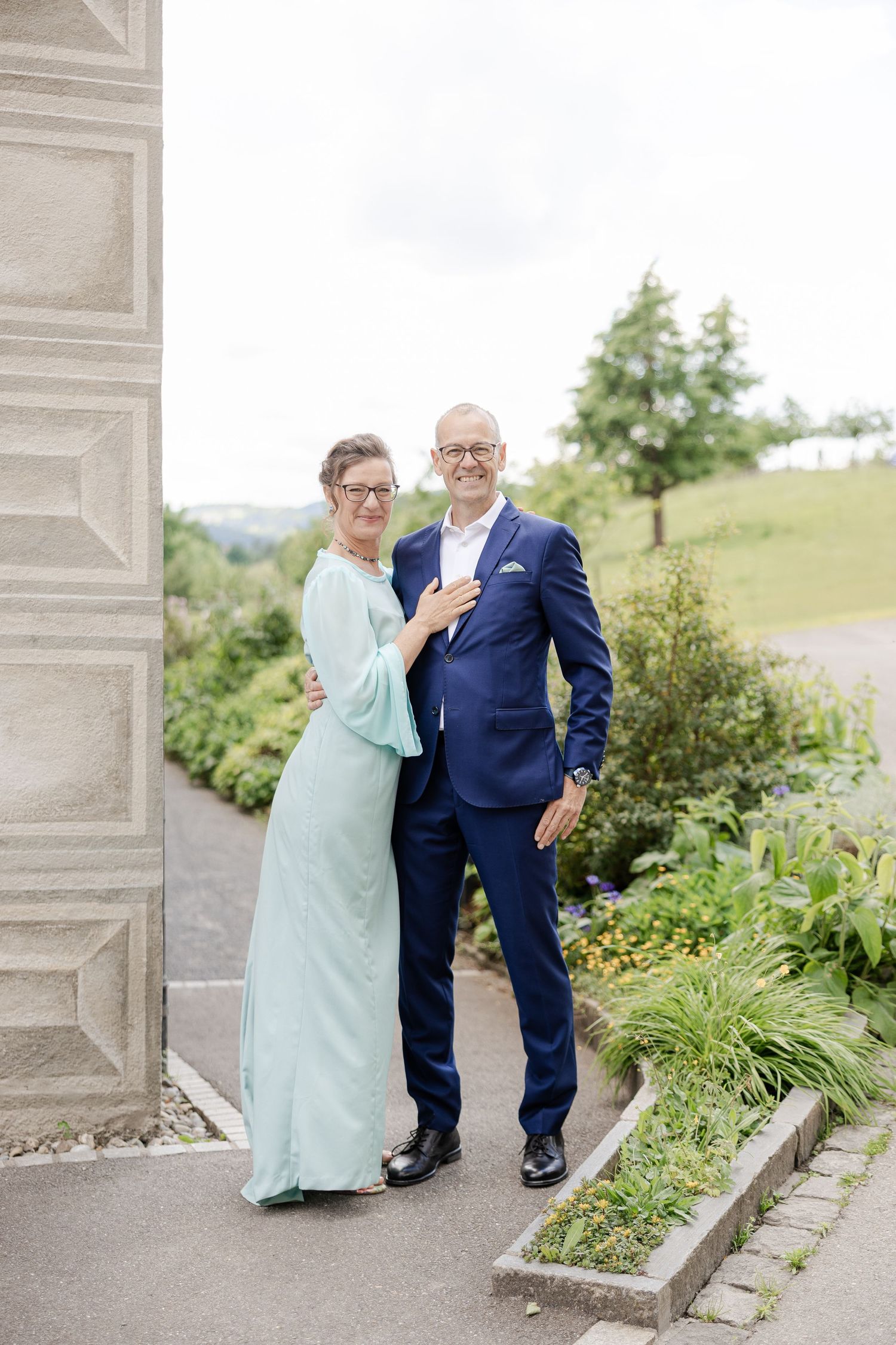 A couple in formal attire poses together outdoors in a garden setting with stone architecture elements - Hochzeitsfotograf Aargau