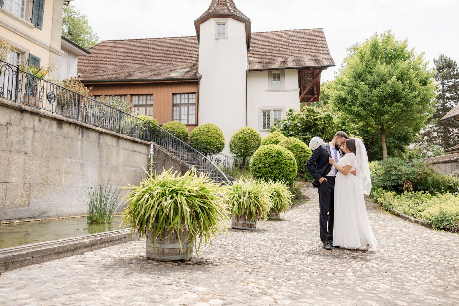 Bride and groom share a romantic moment in an elegant European garden courtyard with manicured topiaries and stone architecture.