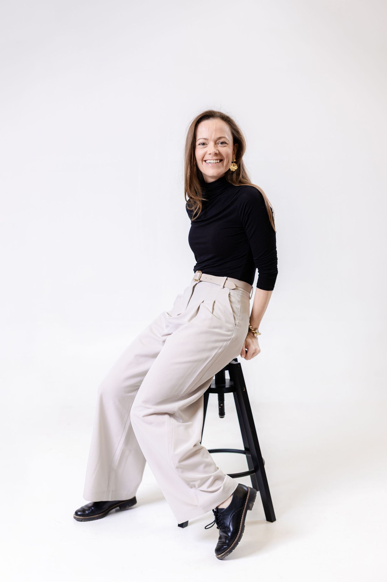 Smiling woman in black turtleneck and beige wide-leg pants sitting on a stool against a white background.