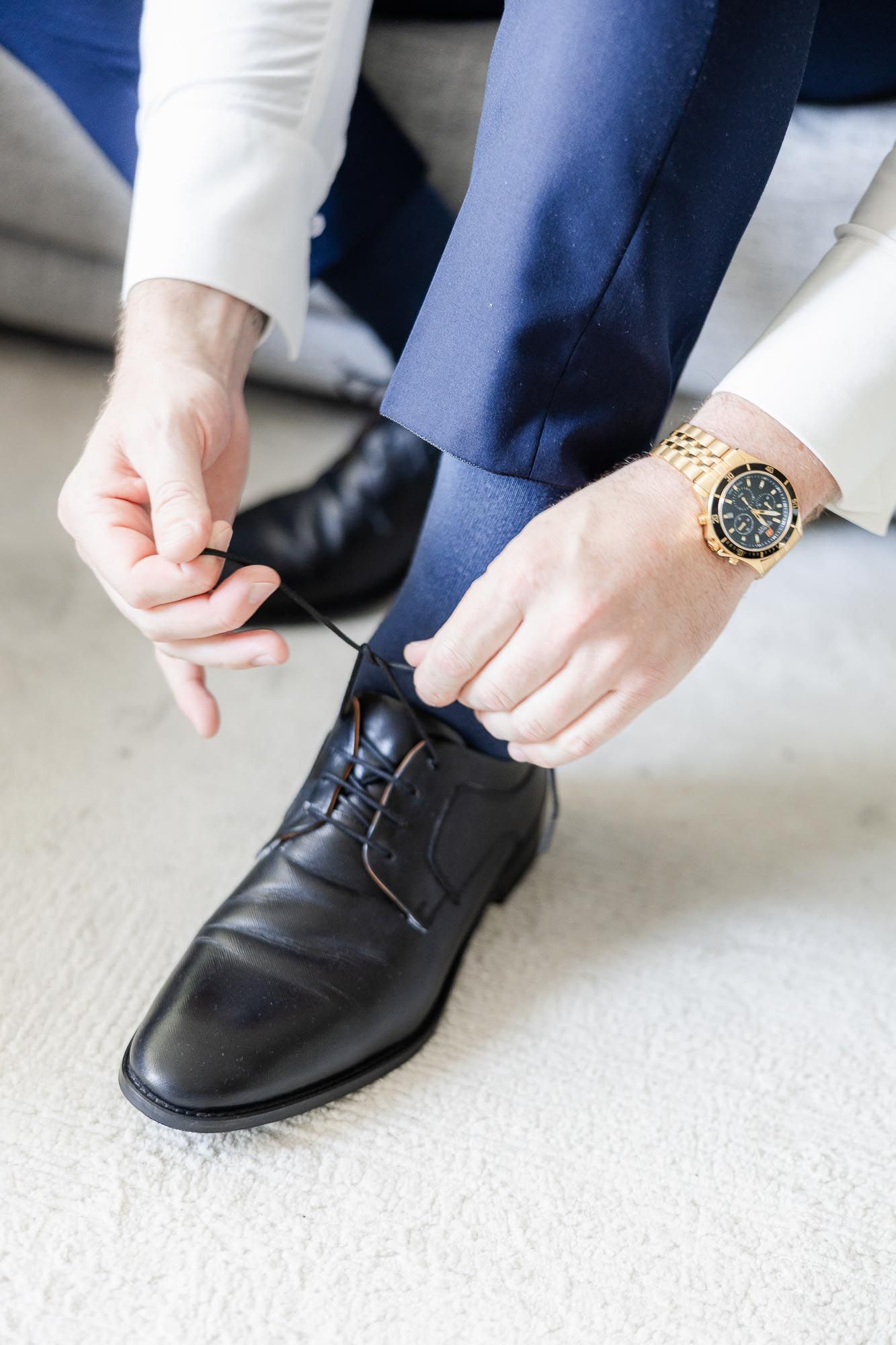 Man in blue suit tying black dress shoes while wearing a gold watch, getting ready for a formal event.