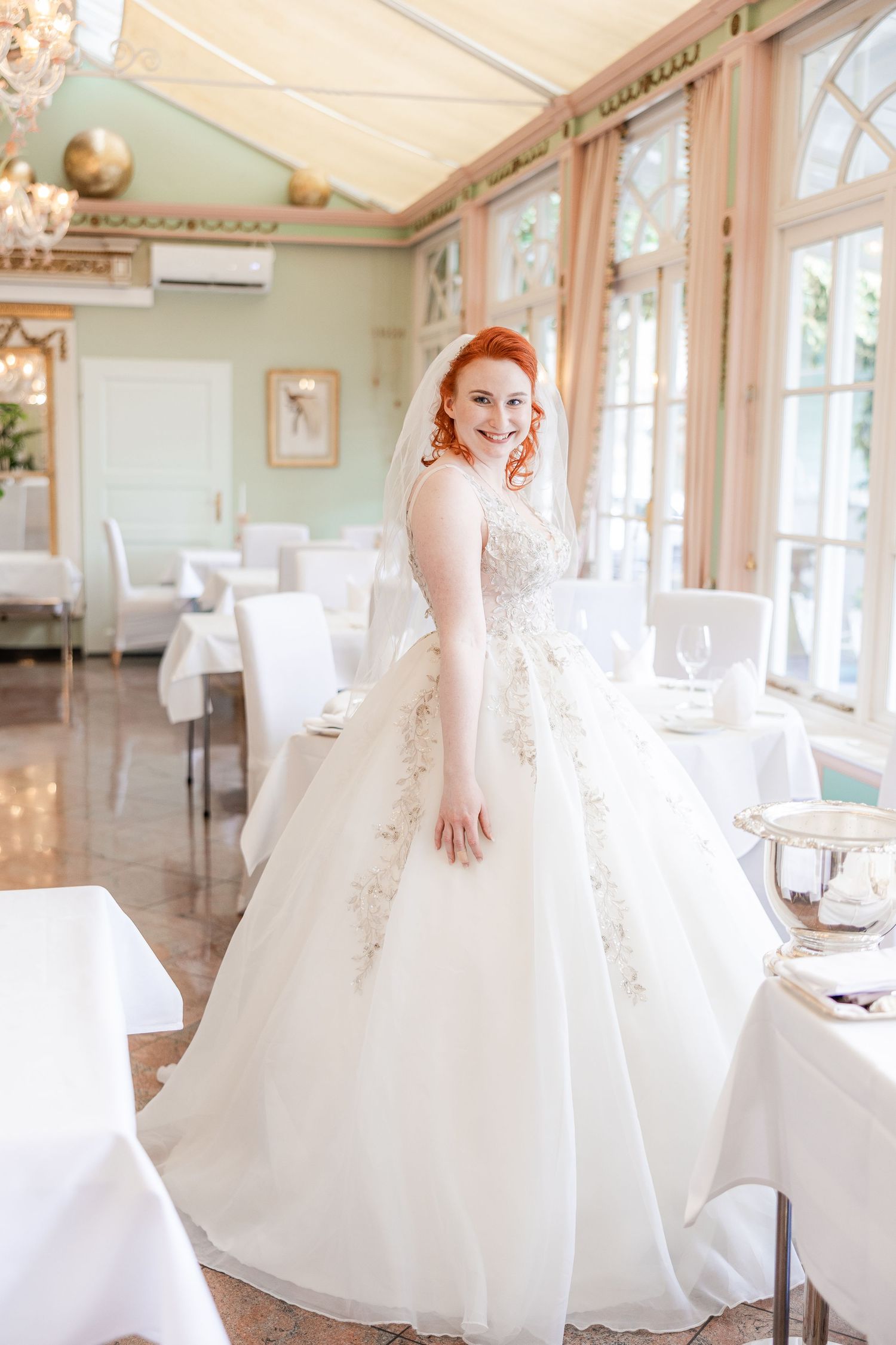 Smiling redhead bride in elegant white ballgown with lace details and veil poses in a sunlit reception hall.