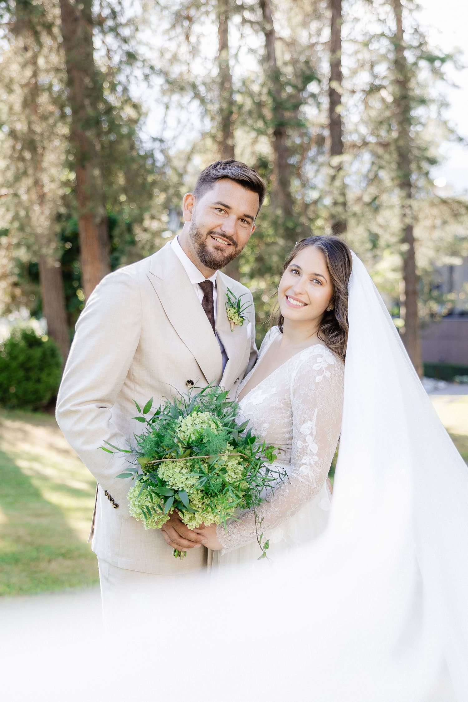 A bride and groom share joyful moments in a wooded outdoor setting with natural greenery and sunlight.