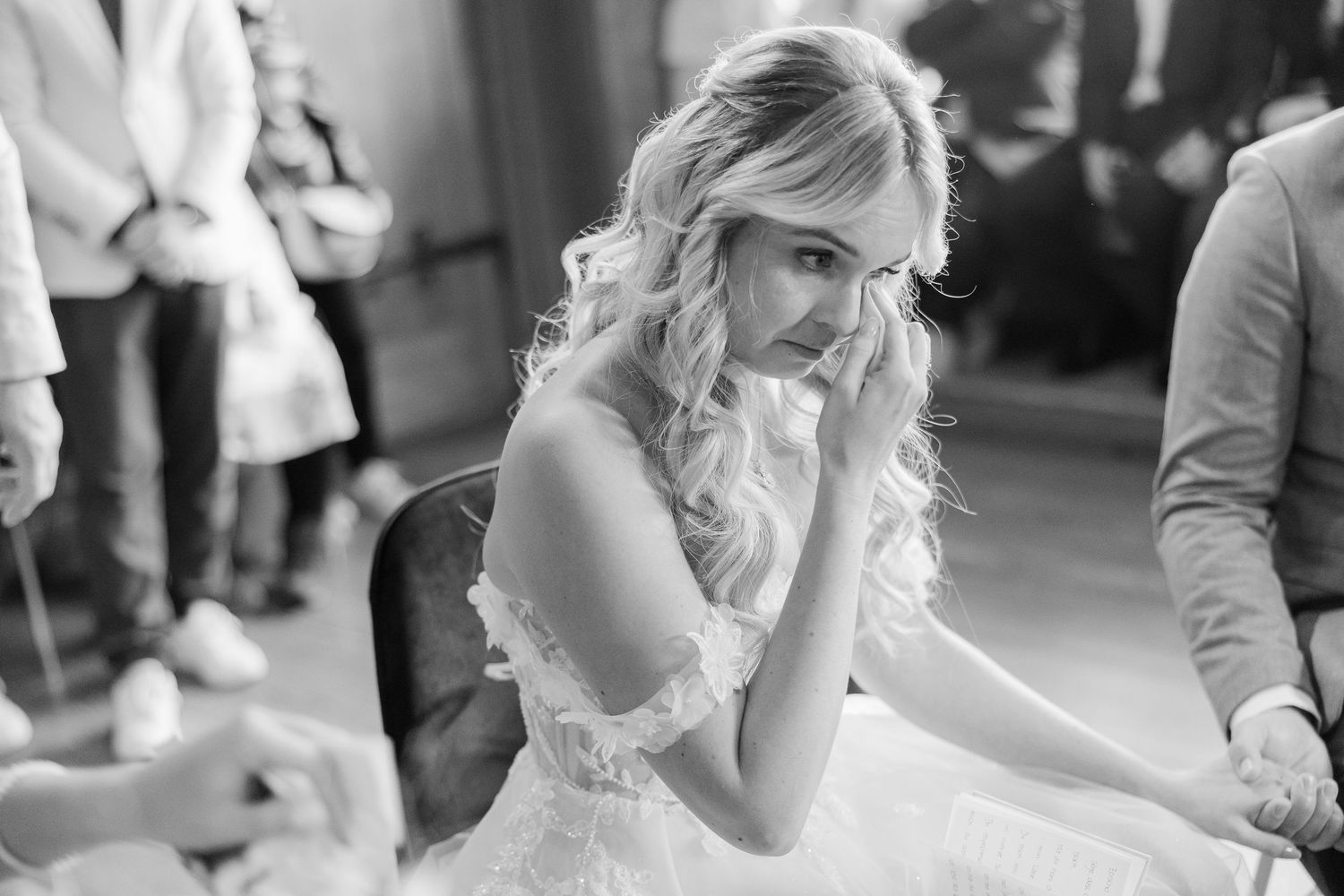 A bride in a lace wedding dress touches her face thoughtfully during preparations in black and white.