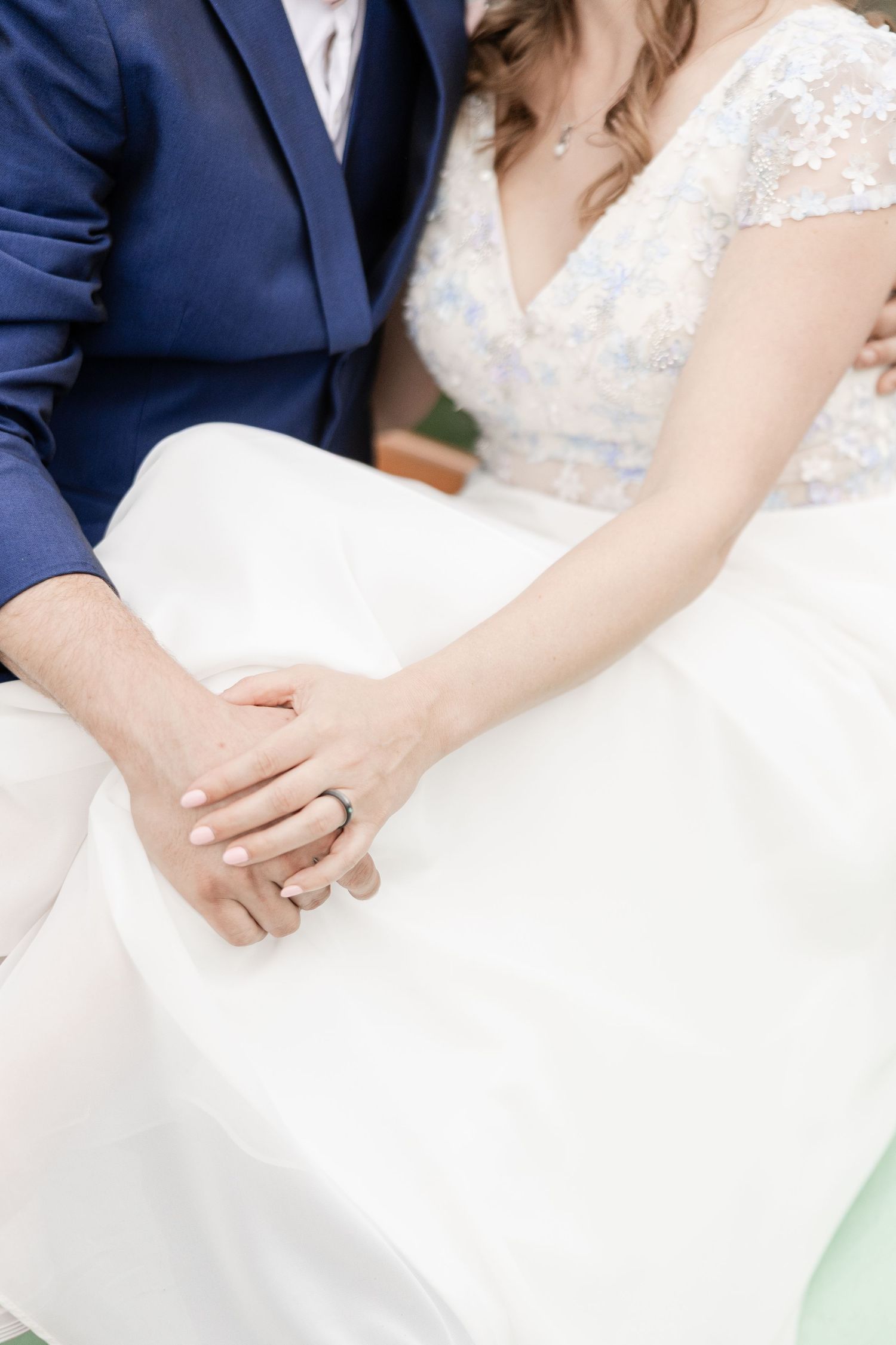 Close-up photo of newlyweds holding hands, with bride in white lace dress and groom in navy suit.