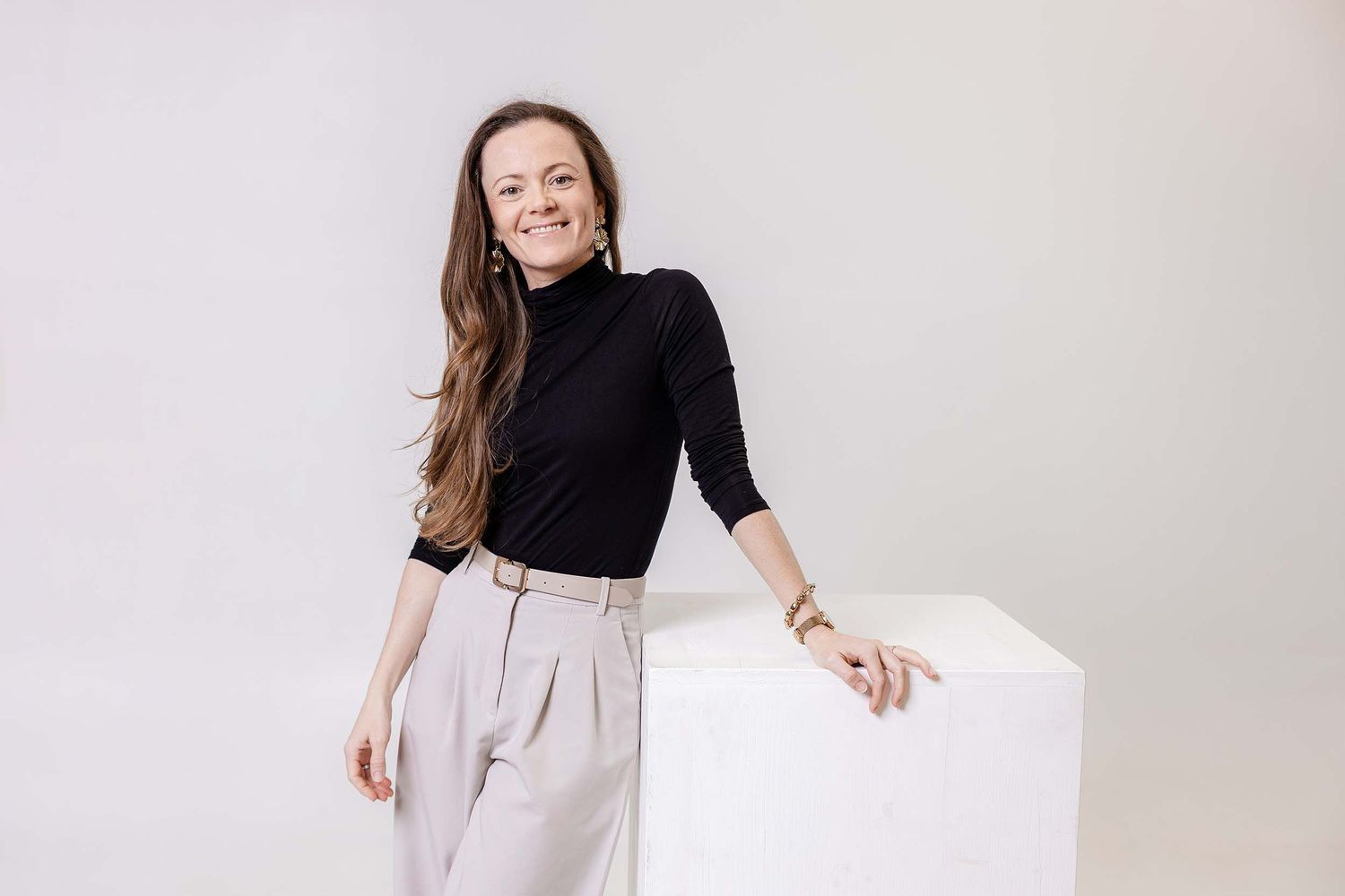 A woman in black turtleneck and beige pants stands confidently against a light background.