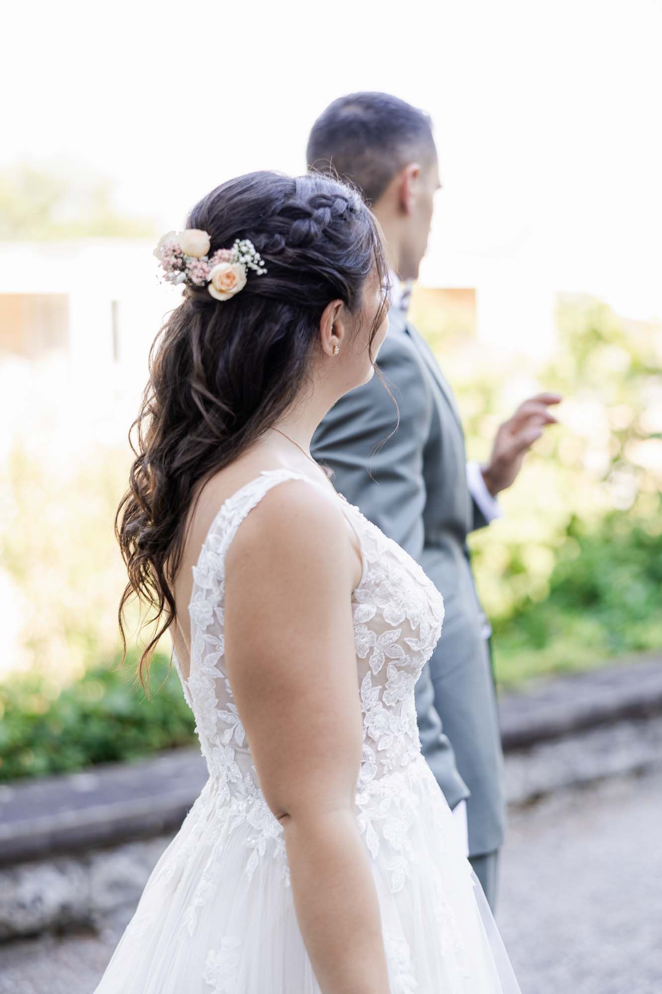 Bride in white lace gown with floral hair accessories stands beside groom in gray suit outdoors.