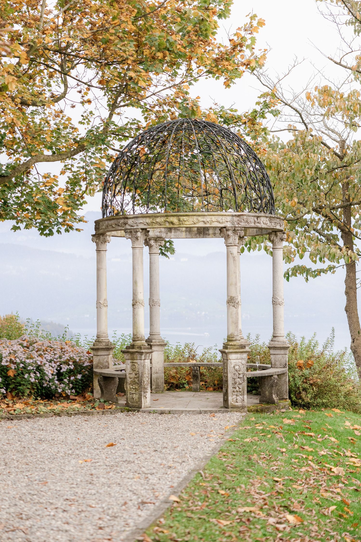 A scenic stone pergola with white columns overlooks a lake, surrounded by autumn trees with golden leaves.