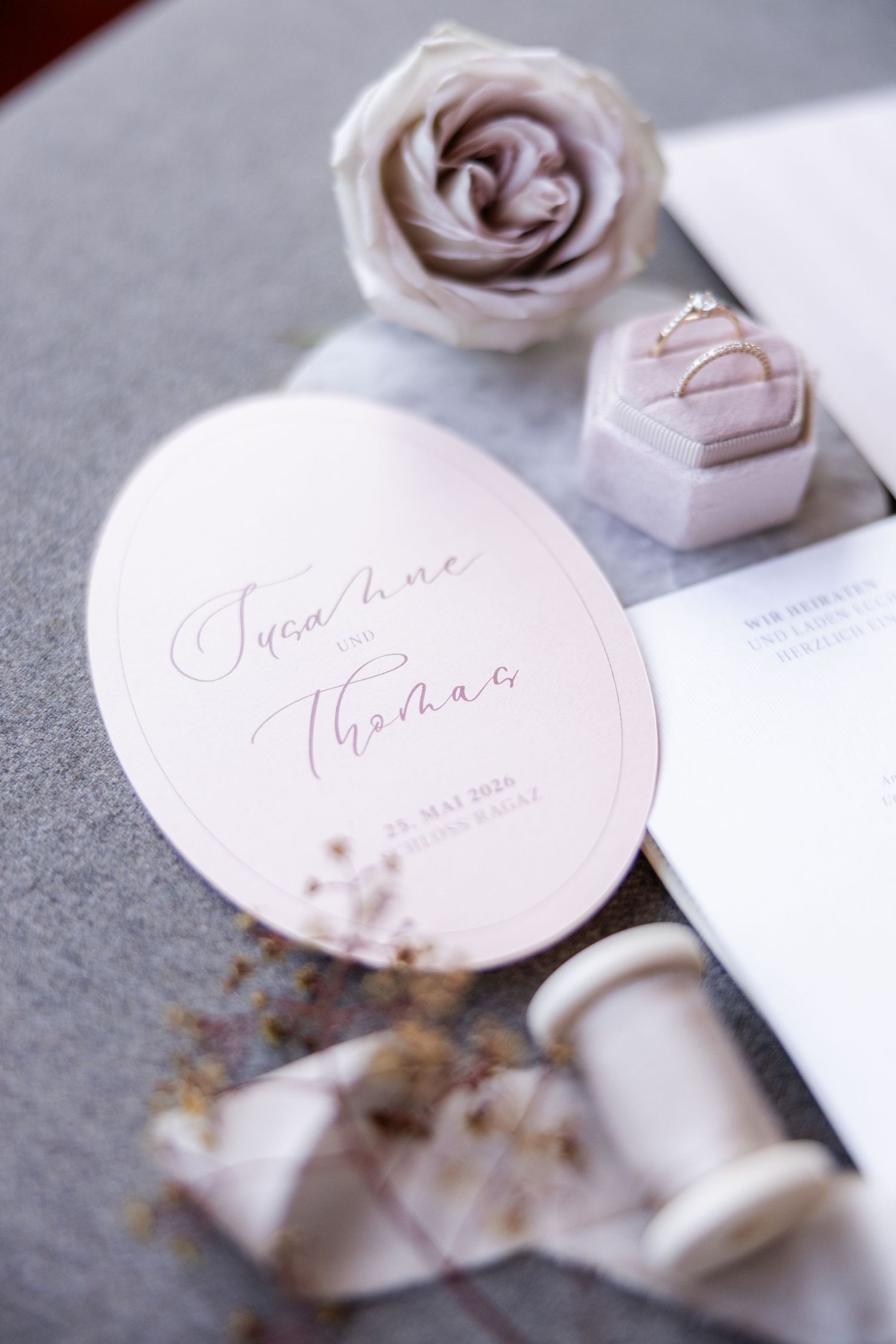 Elegant wedding stationery flat lay with blush oval invite, rings in velvet box, silk ribbon, and dried florals.