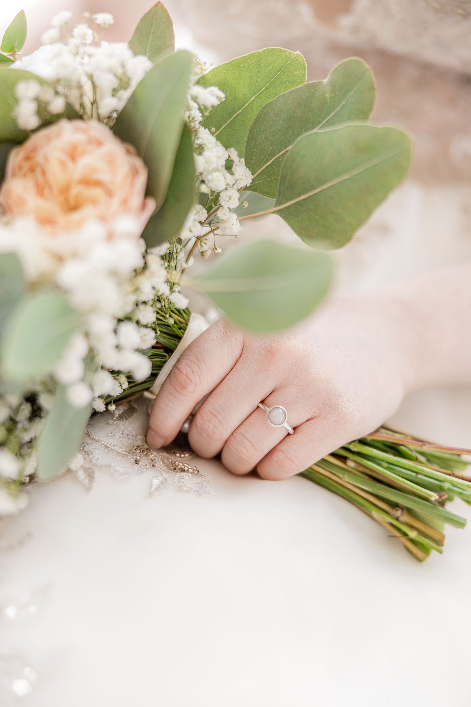 Bride holding a wedding bouquet with peach roses, baby's breath, and eucalyptus, wearing a delicate diamond engagement ring.