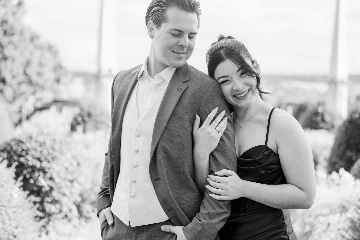 Black and white photo of smiling engaged couple, woman showing ring, man in suit, outdoors.