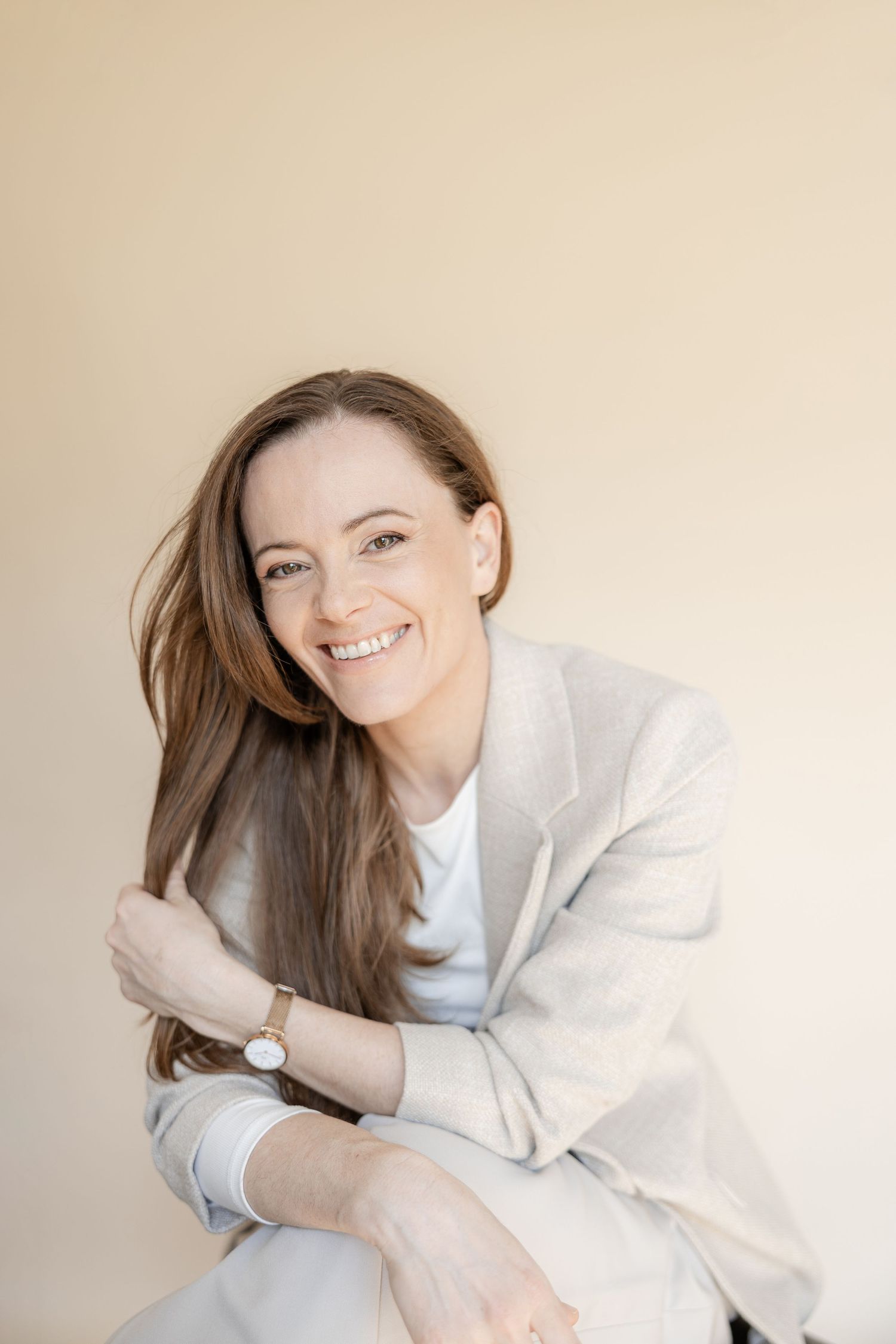 Smiling woman with long brown hair wearing a light beige blazer and watch, posing against a neutral background.