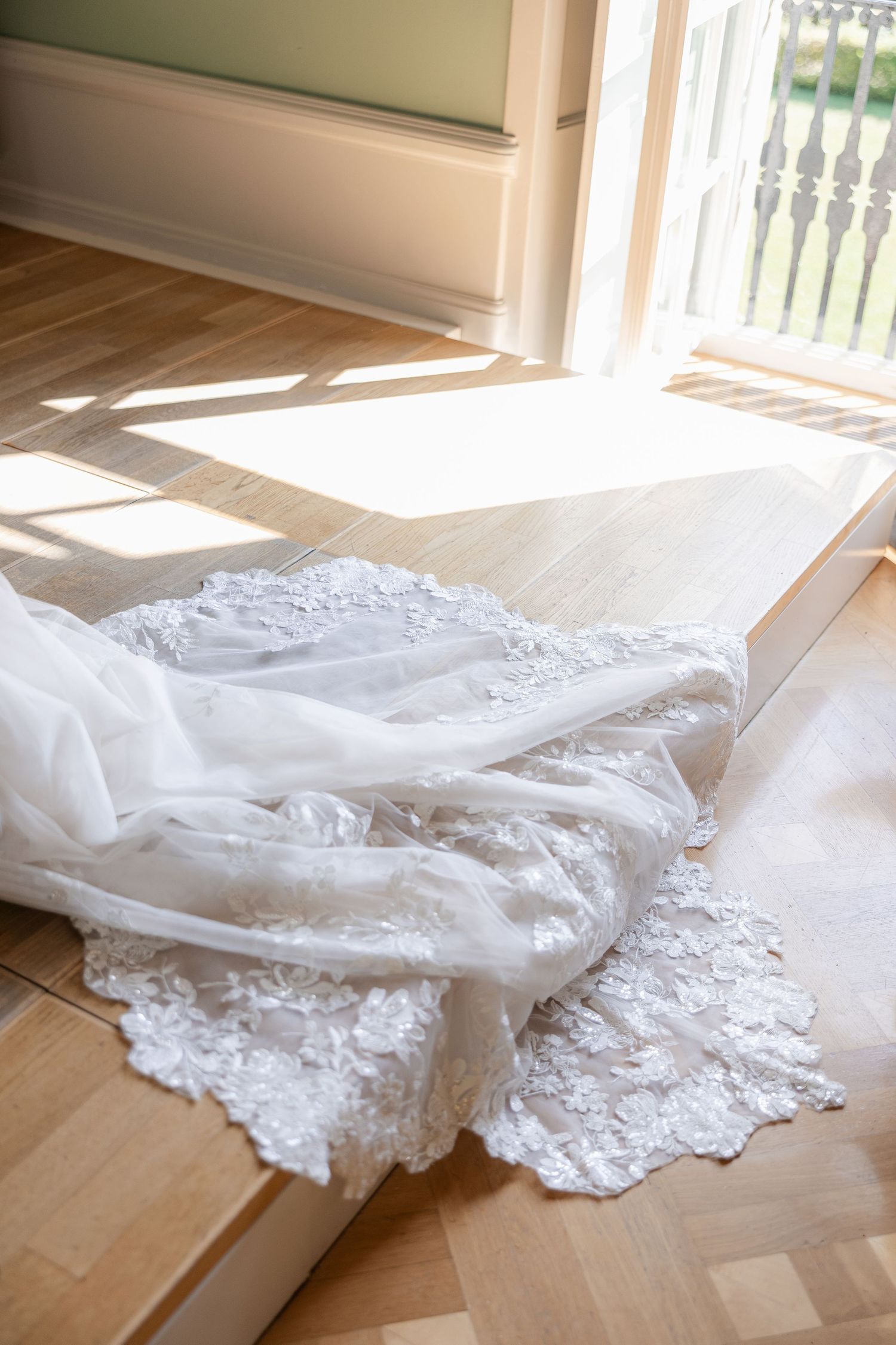White lace wedding dress train spread on a sunlit wooden floor near a bright window.