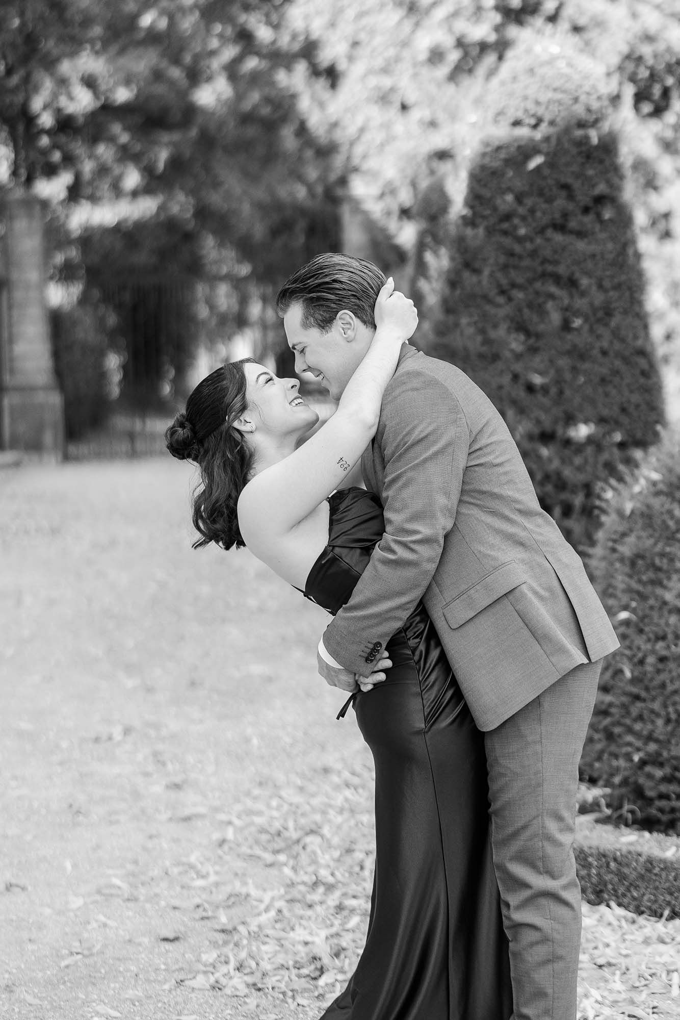 Black and white photo of a couple sharing a romantic dip kiss outdoors in a garden setting.