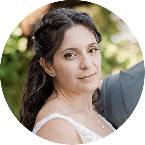 A bride in a white lace dress smiles softly outdoors, with dark wavy hair adorned with a delicate hair accessory.