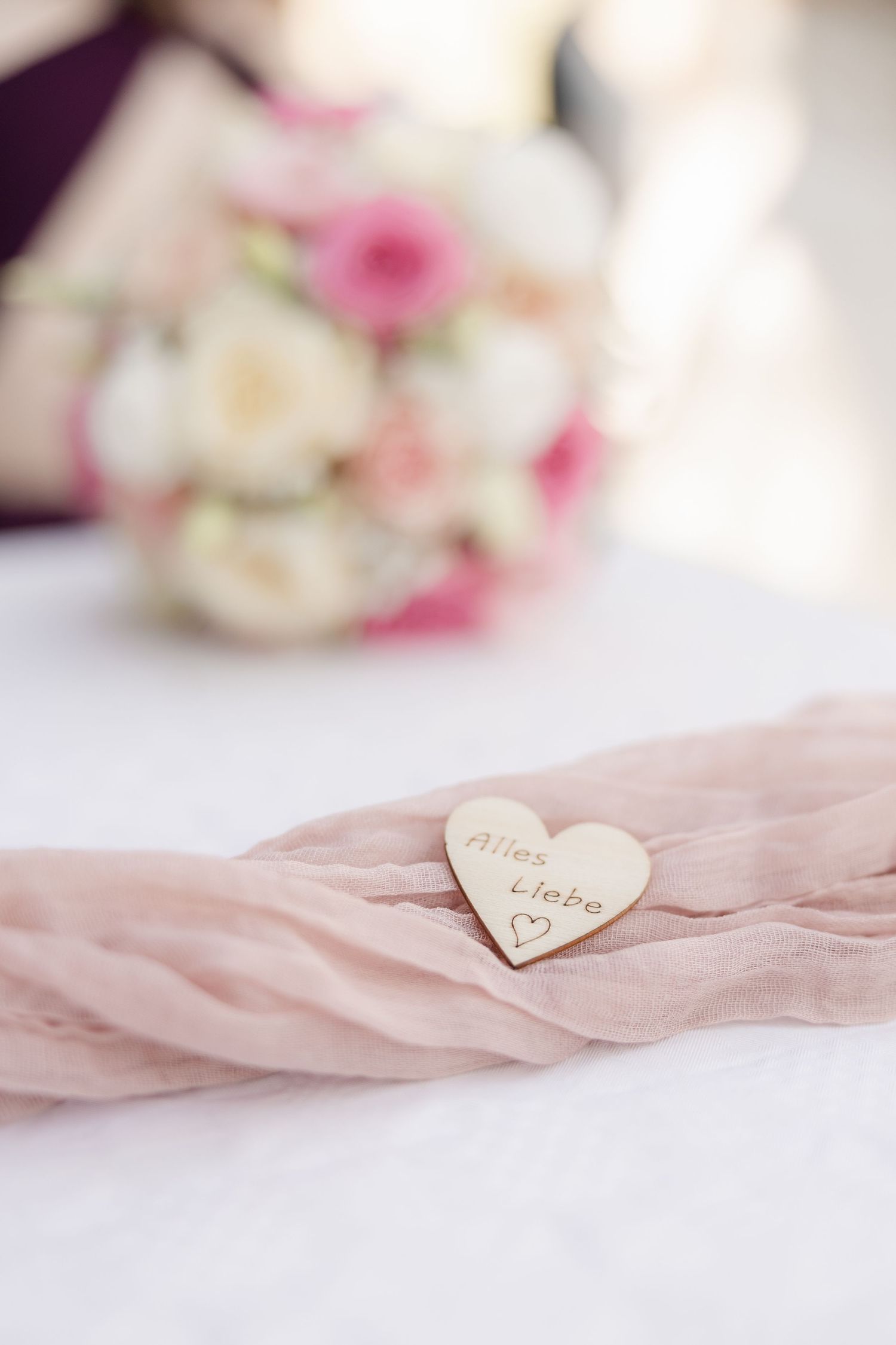 A wooden heart decoration on pink fabric with a romantic blurred flower bouquet in the background.