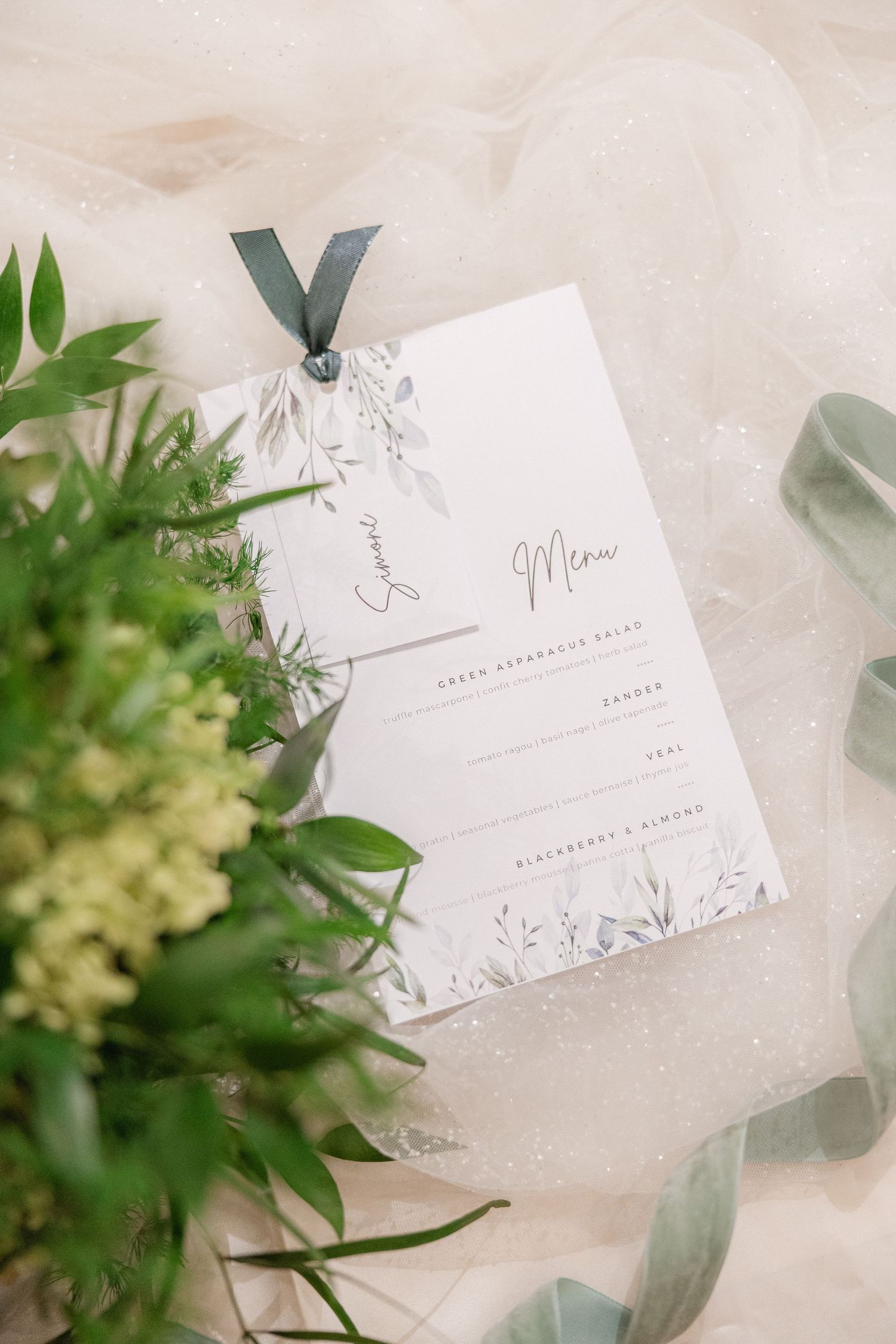 Elegant wedding menu card with delicate floral details and gray ribbon displayed among greenery and white flowers.