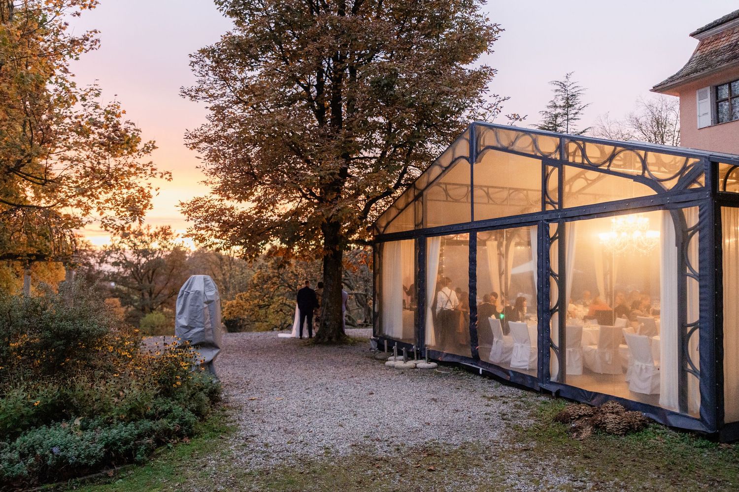 A modern glass-walled building glows warmly at sunset with trees and natural landscaping surrounding the structure.