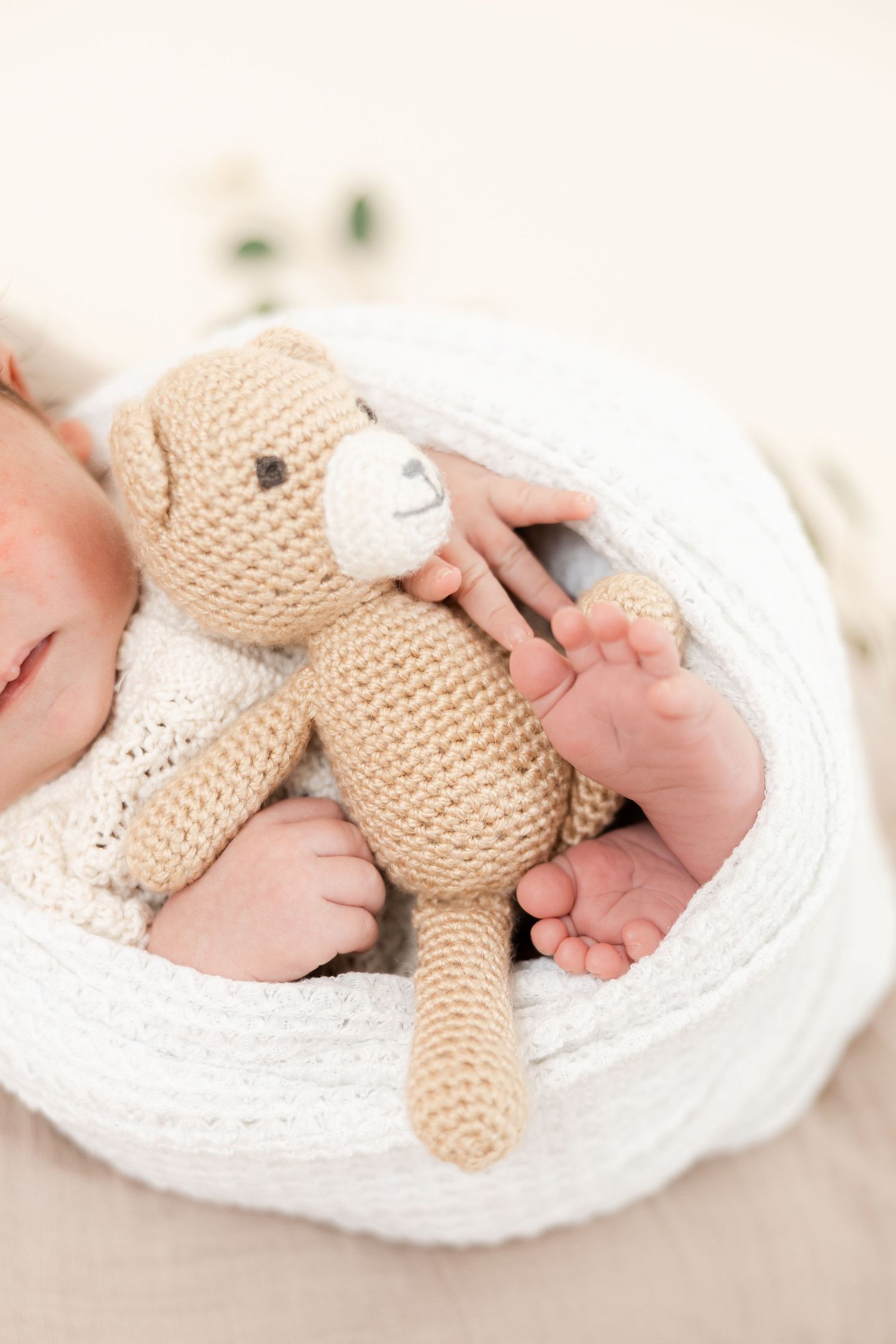 A tiny baby's feet resting on a white blanket with a soft knitted beige teddy bear companion.