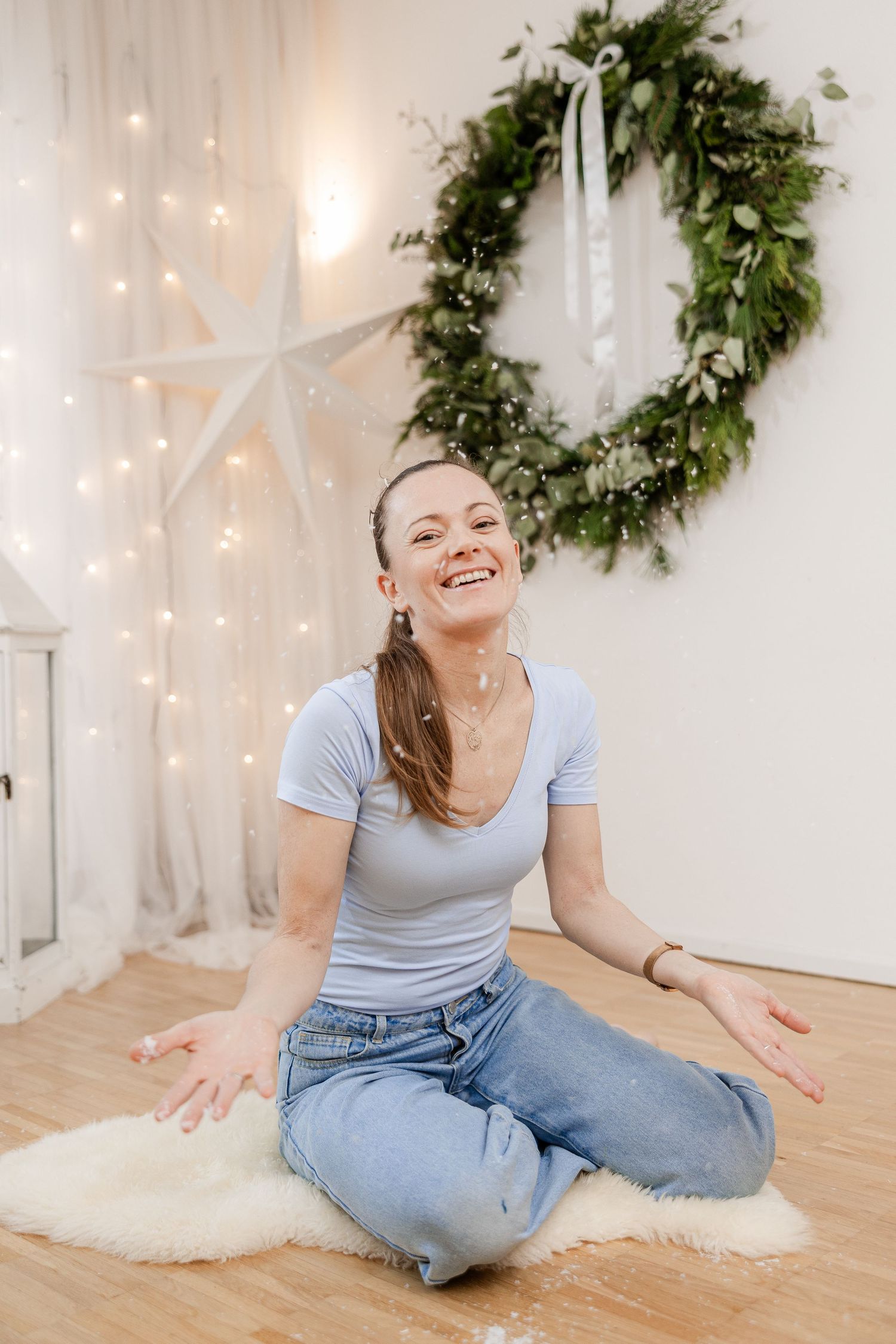 Woman in light gray t-shirt and jeans sits on wooden floor with holiday decor and twinkling lights in background.