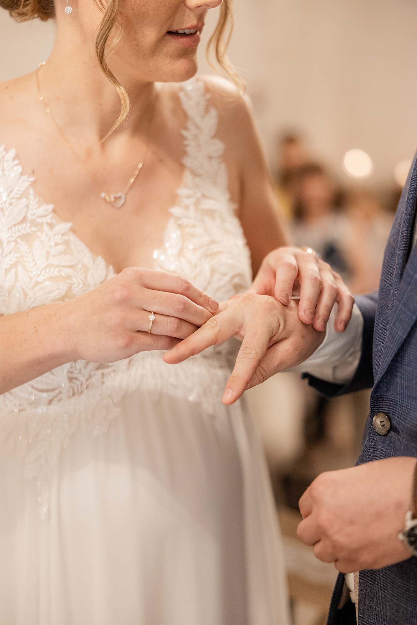 Bride placing a wedding ring on groom's finger during ceremony, wearing lace gown and diamond necklace.