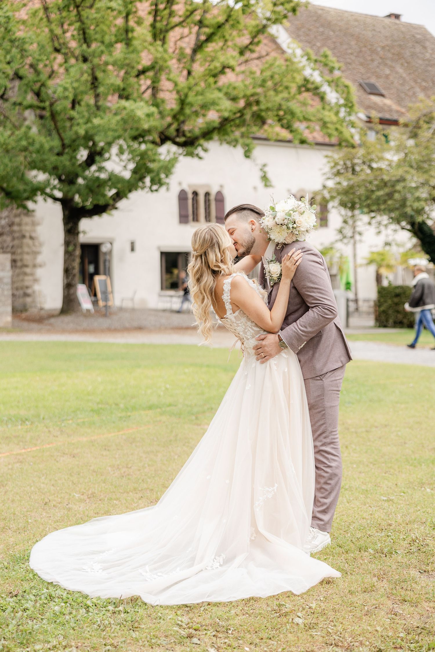 A romantic wedding couple embraces and kisses in an outdoor garden setting with a flowing white dress and grey suit.