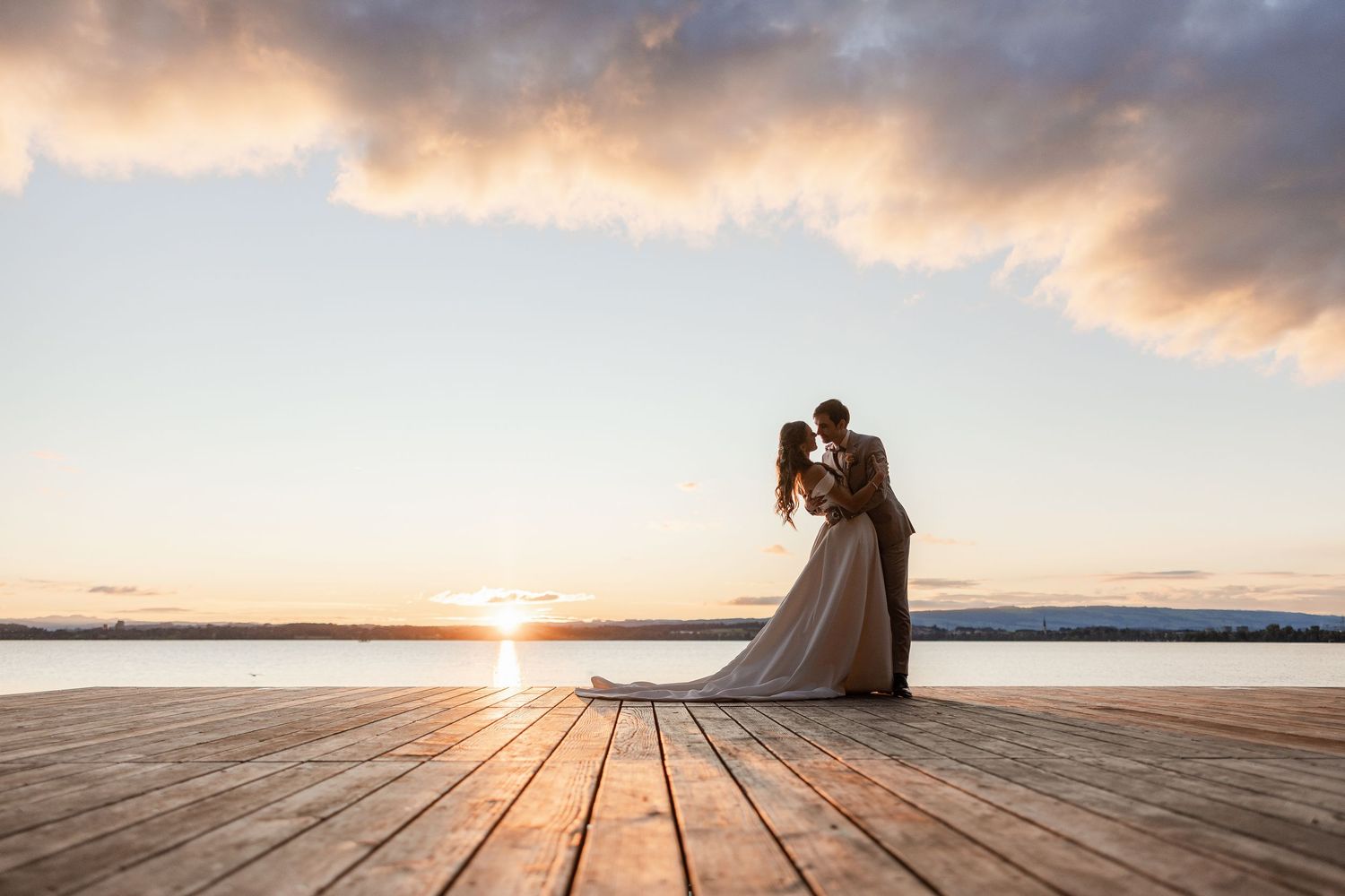 Silhouettes of couple embracing on wooden pier at sunset over tranquil lake.
