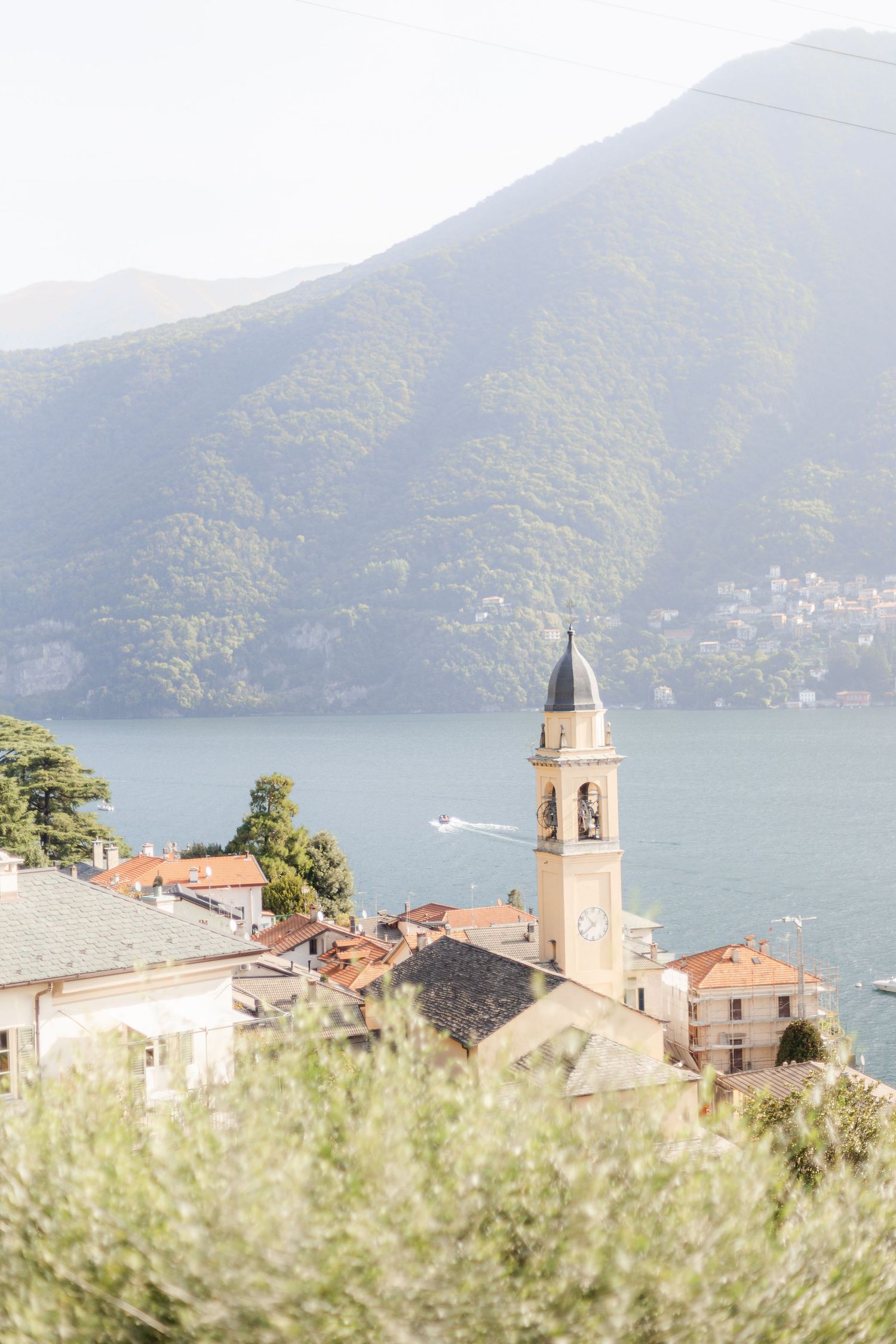 Church bell tower rises above terracotta roofs along Lake Como in Italy with misty mountains in the background.