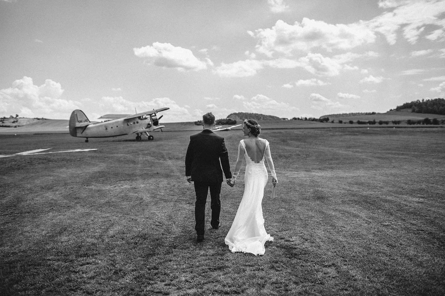 A couple walks across an open field with vintage airplanes parked in the distance on a cloudy day.