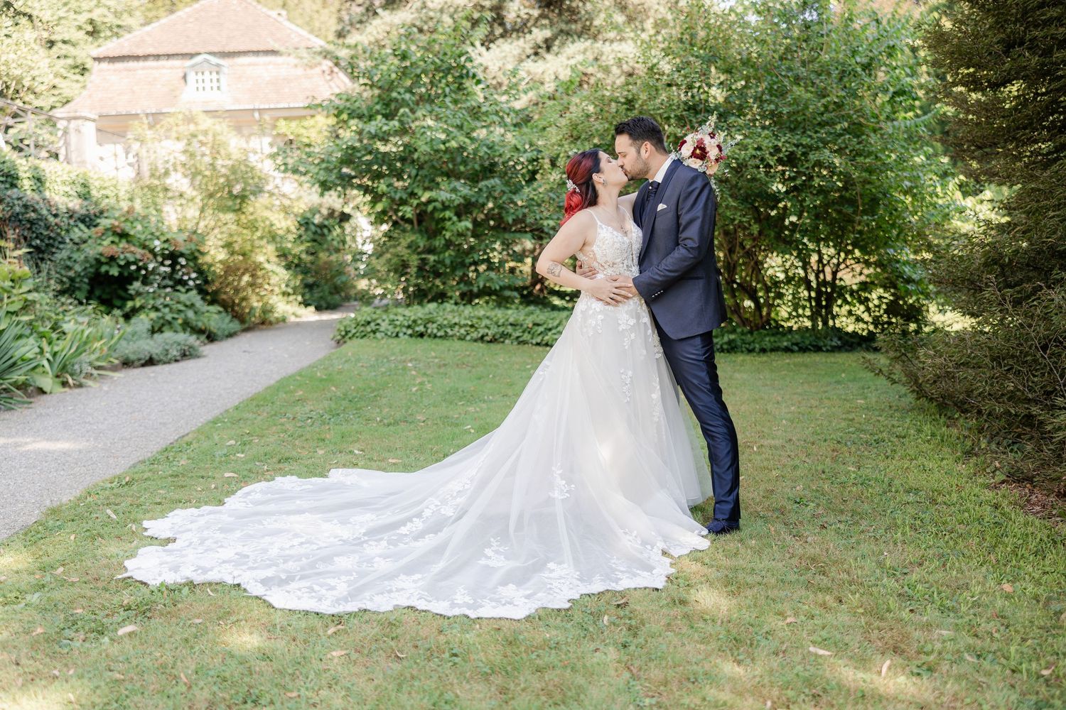 A couple in wedding attire embraces on a garden path with a flowing white dress train.
