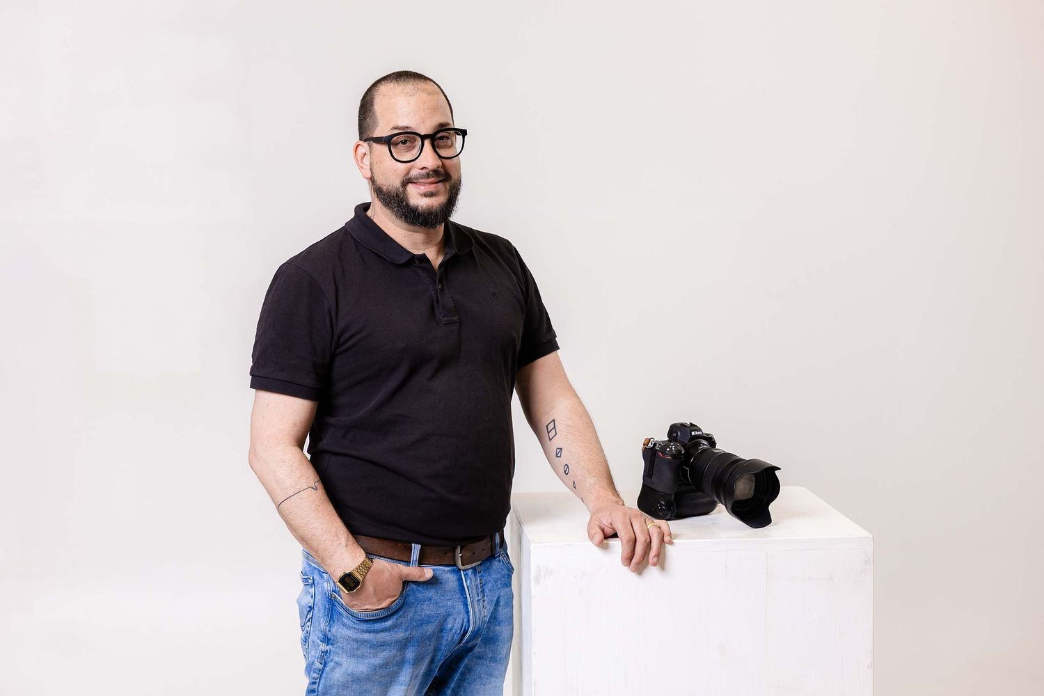 Professional photographer in black polo shirt and jeans stands next to camera equipment against white studio backdrop.