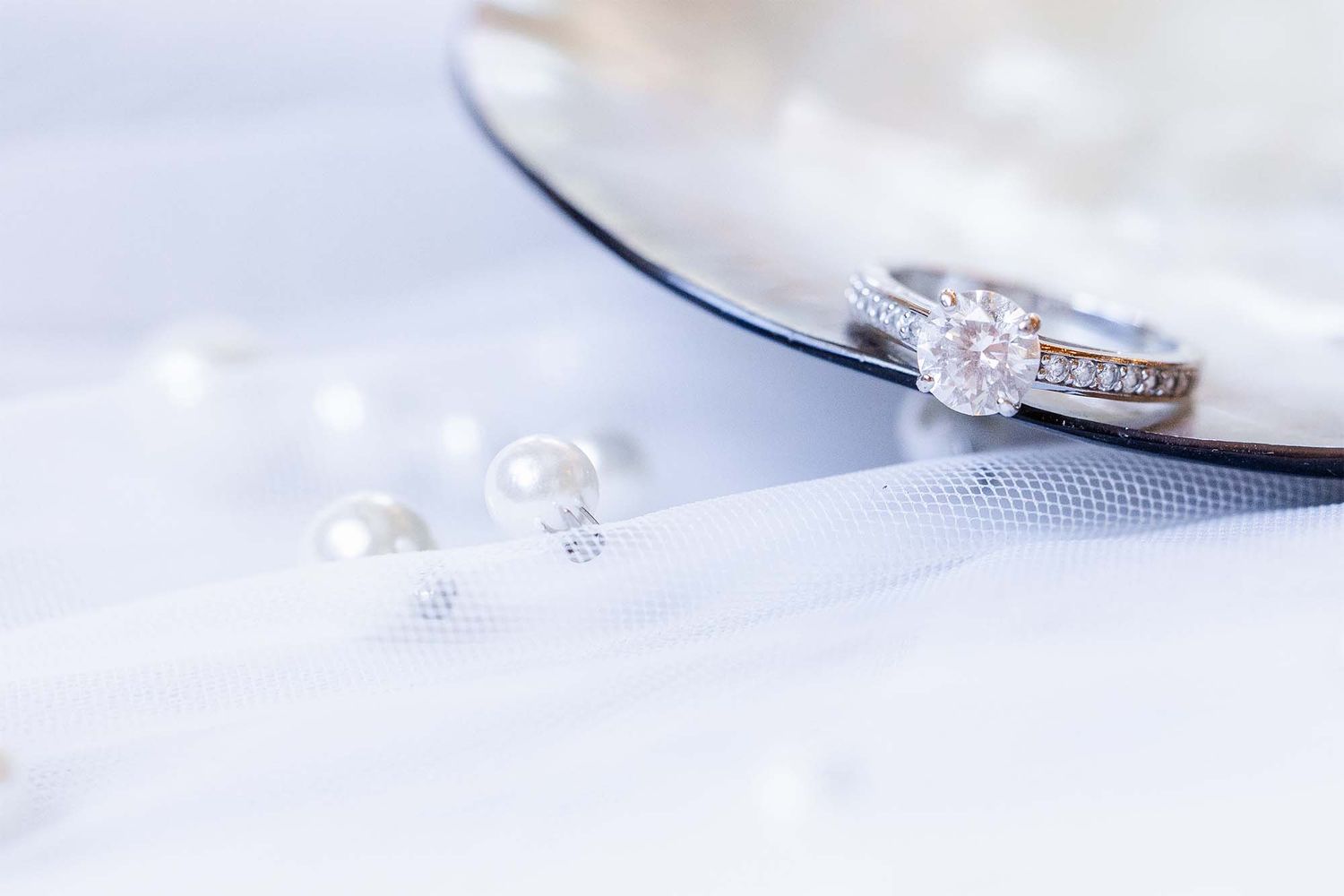 Elegant diamond engagement ring and wedding band displayed with pearl earrings on soft white fabric.
