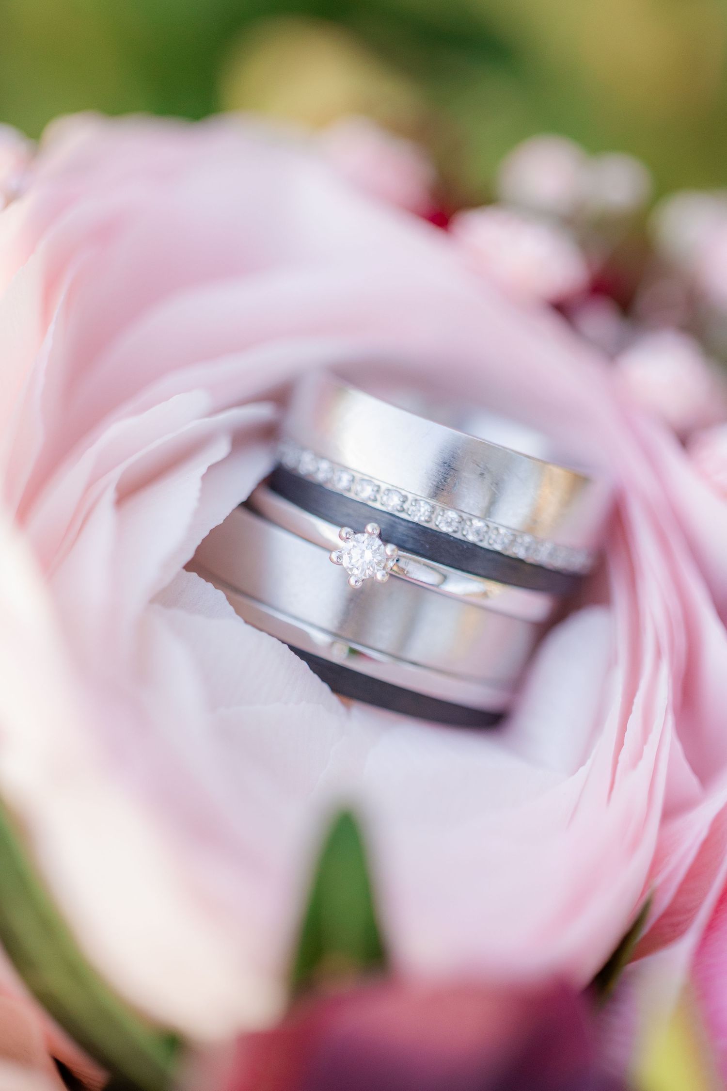 Three silver wedding rings with diamonds nestled in soft pink ranunculus flowers in a bridal bouquet.