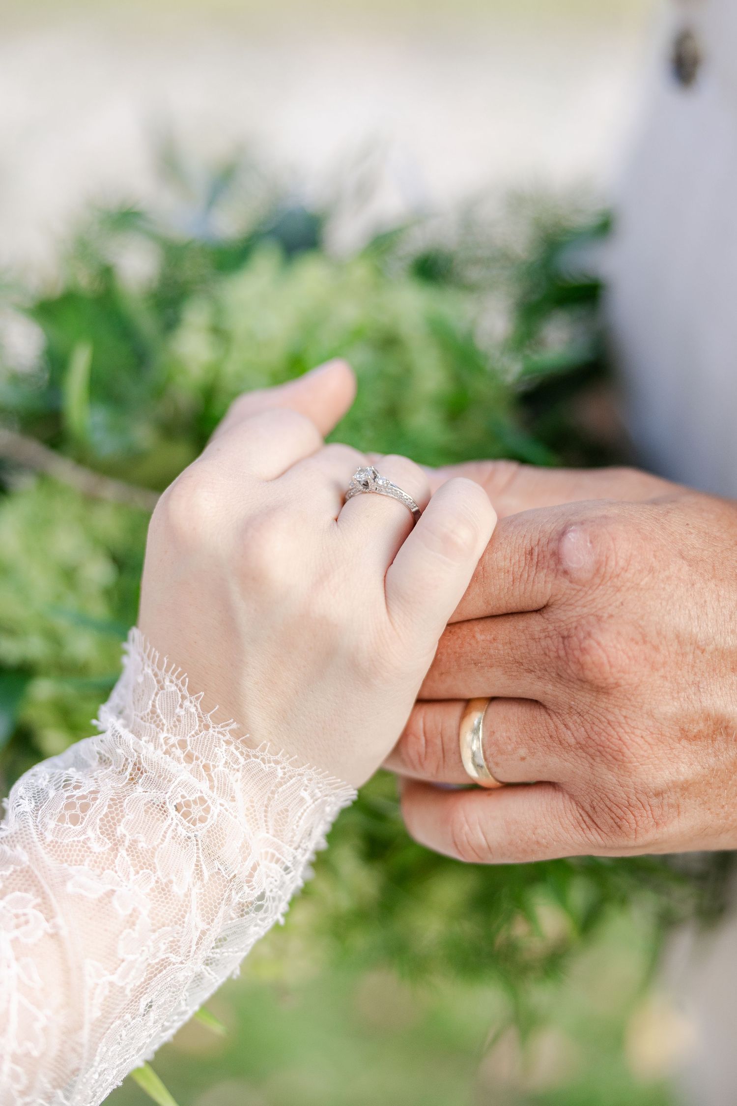 Close-up view of hands with wedding rings against a soft green outdoor background showing delicate white lace details.