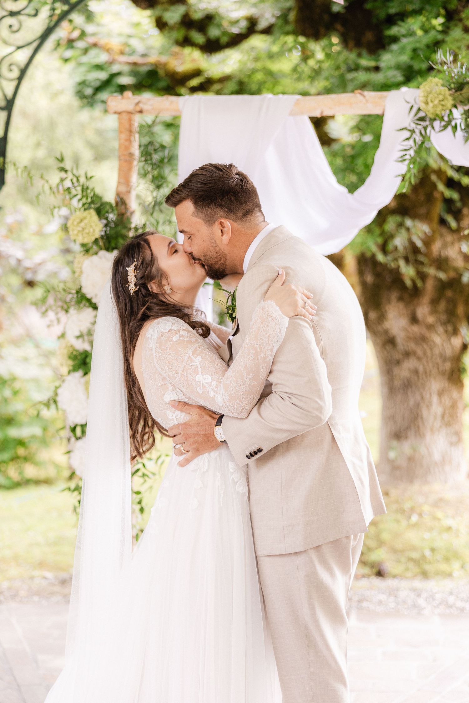 A romantic wedding moment captured under a white draped arch with floral decorations in a garden setting.