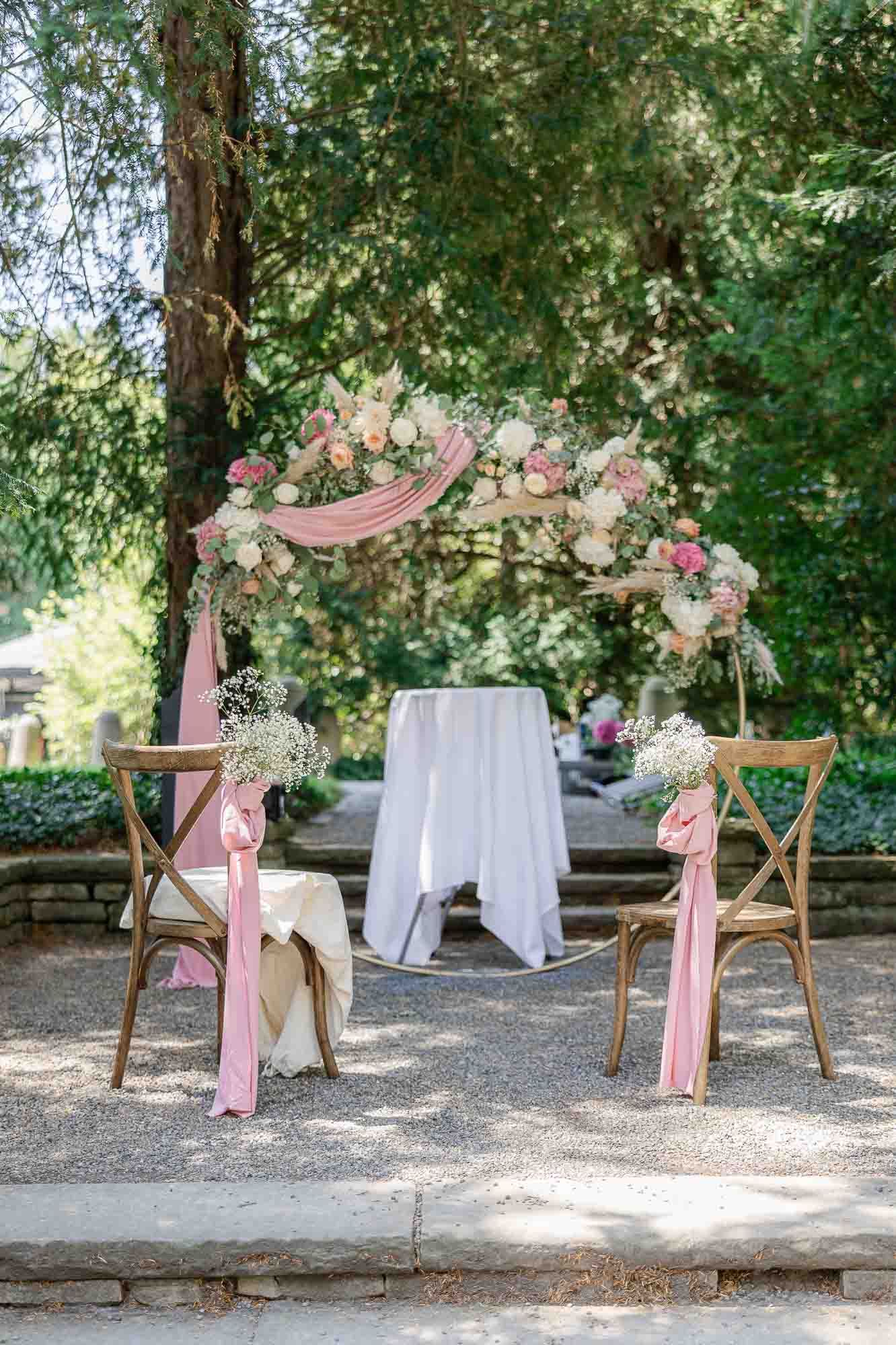 Elegant outdoor wedding arch with pink draping, floral arrangements, and wooden cross-back chairs set in a lush garden setting.