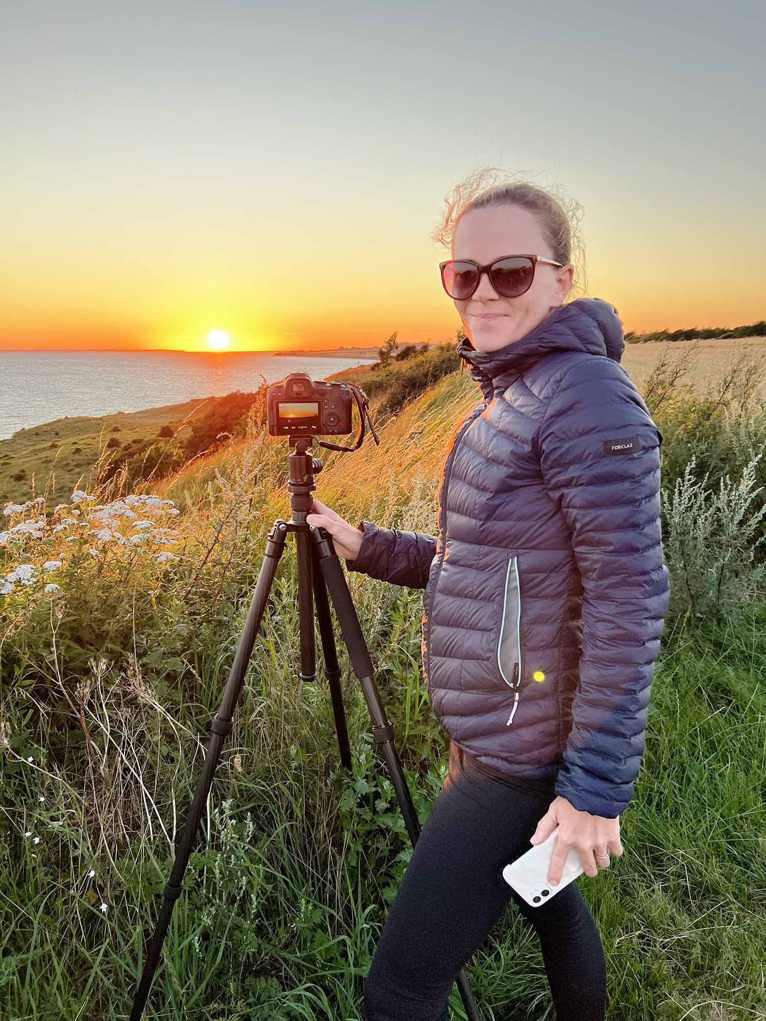 Photographer with camera on tripod captures stunning coastal sunset over water while standing on grassy cliff.