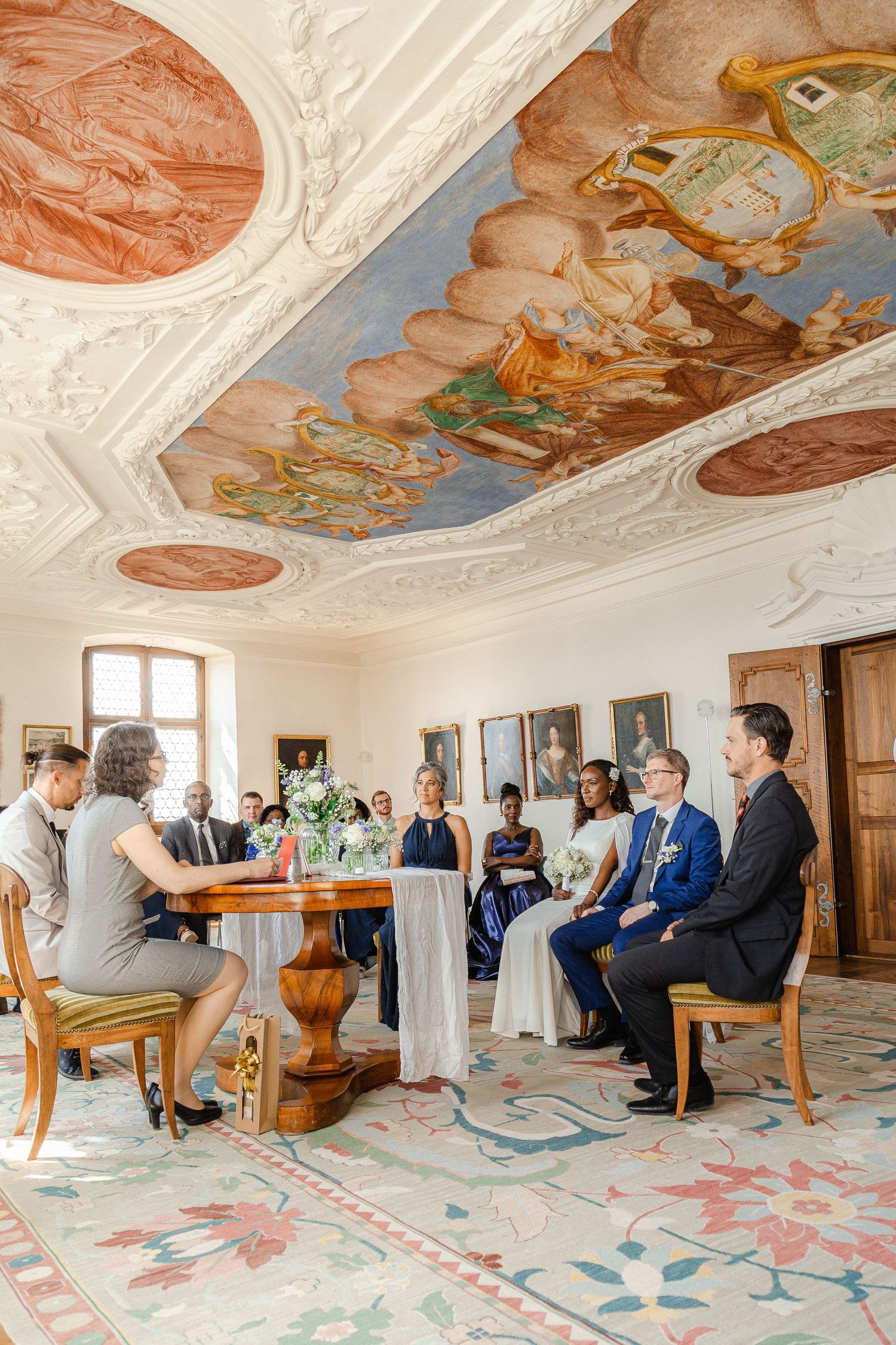 Ornate baroque ceiling fresco adorns a room where a small group gathers around a wooden table on patterned carpet.