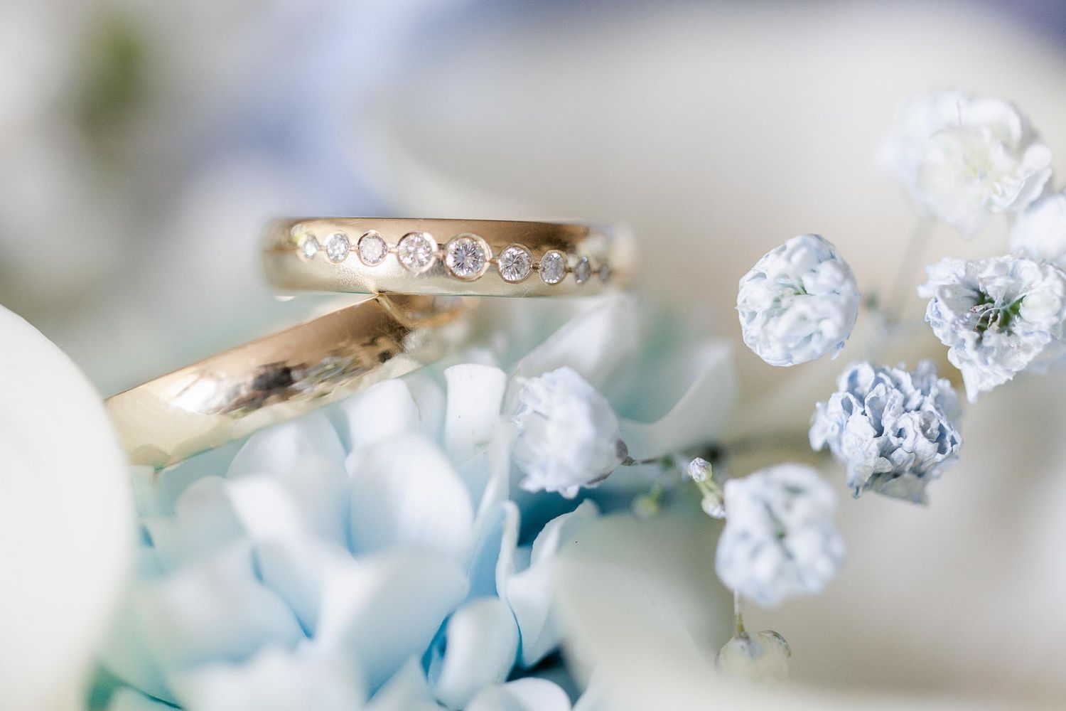 Two gold wedding rings with diamonds rest on blue hydrangea flowers and white baby's breath in a soft bokeh setting.