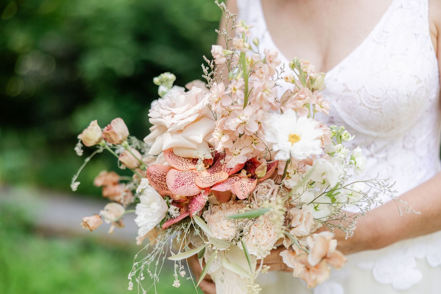 Bride holding a romantic bridal bouquet with peach roses, white daisies, and wildflowers in a lush garden setting.