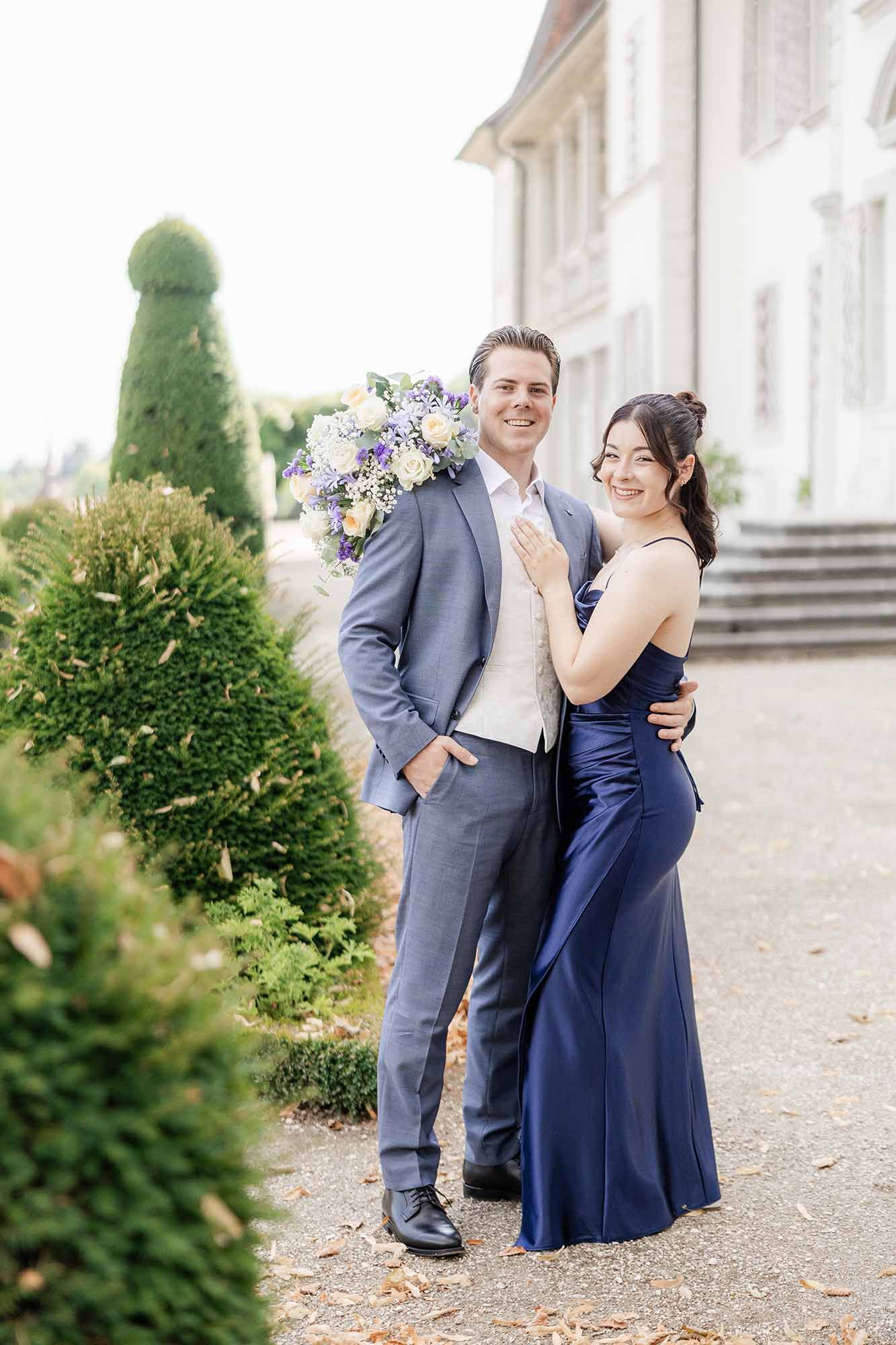 Couple posing outdoors, man in gray suit holding bouquet, woman in navy blue gown, elegant garden setting.