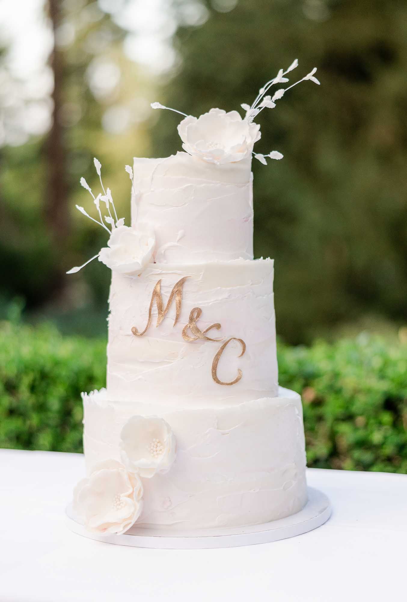 Three-tier white wedding cake with gold M&C monogram, white sugar flowers, and textured frosting displayed outdoors.
