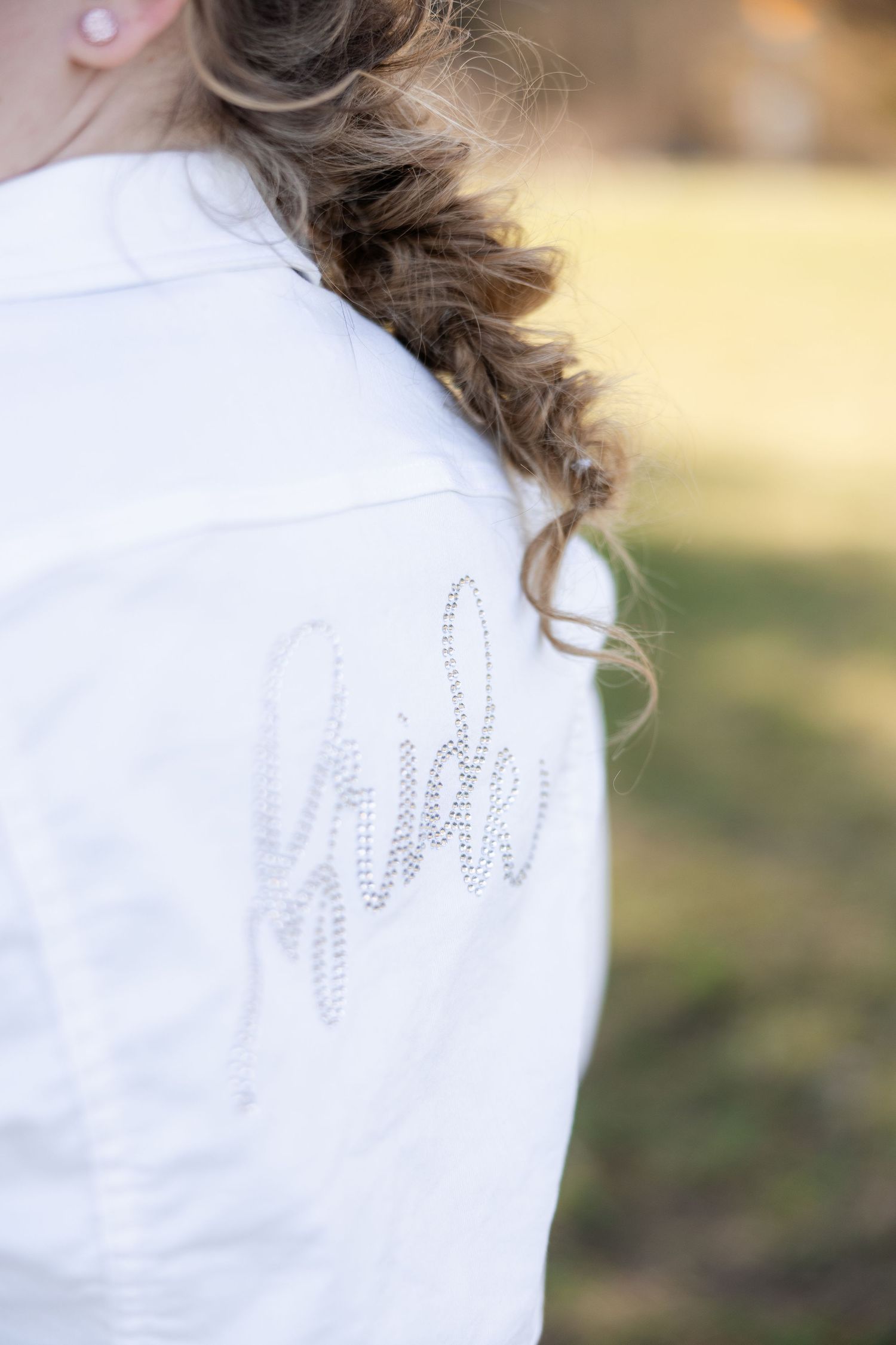 Close-up of a white bridal jacket with rhinestone 'Bride' script on the back, worn by a woman with a braided hairstyle.