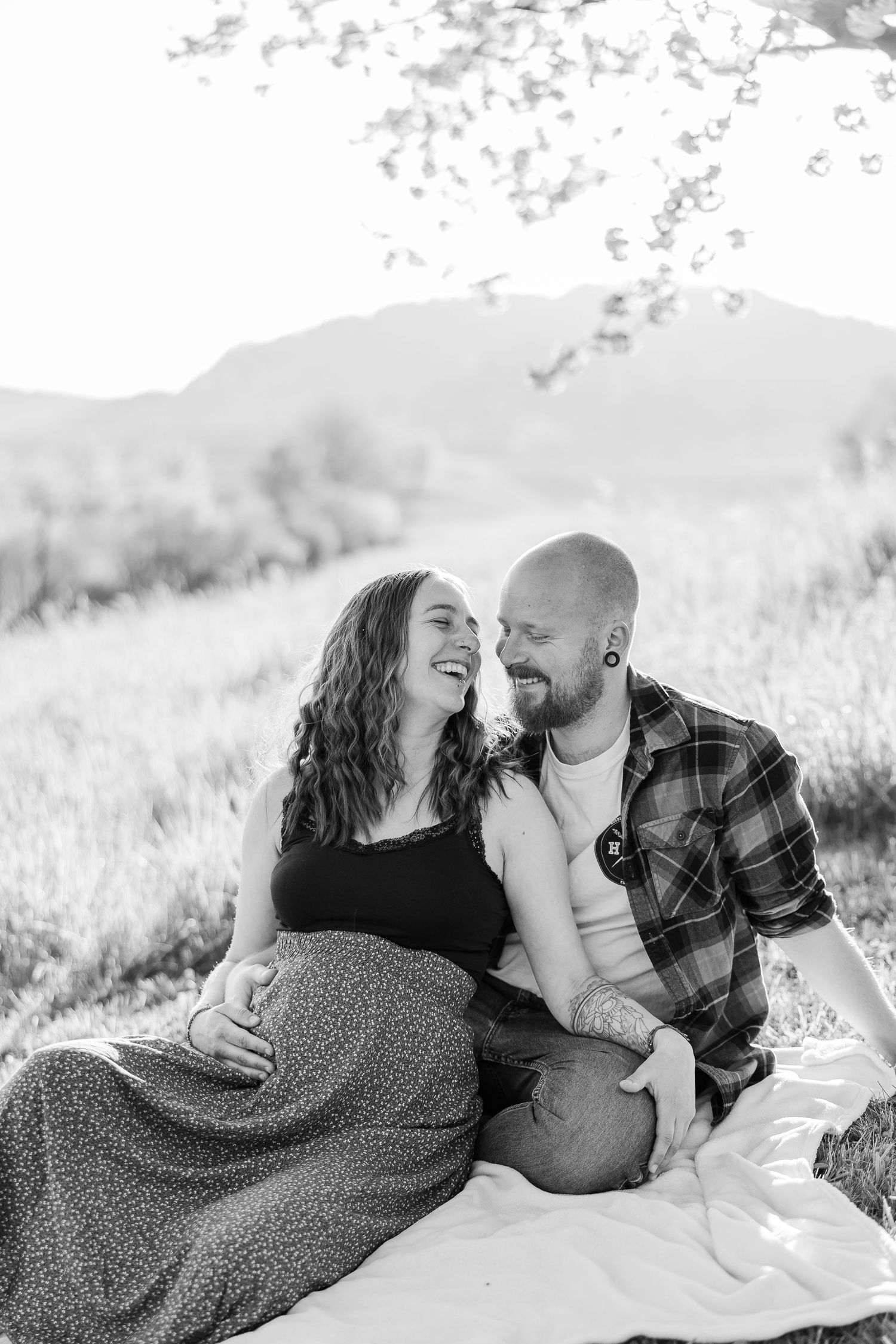 A couple shares a joyful moment together while sitting on a blanket in a sunny meadow with mountains in the background.