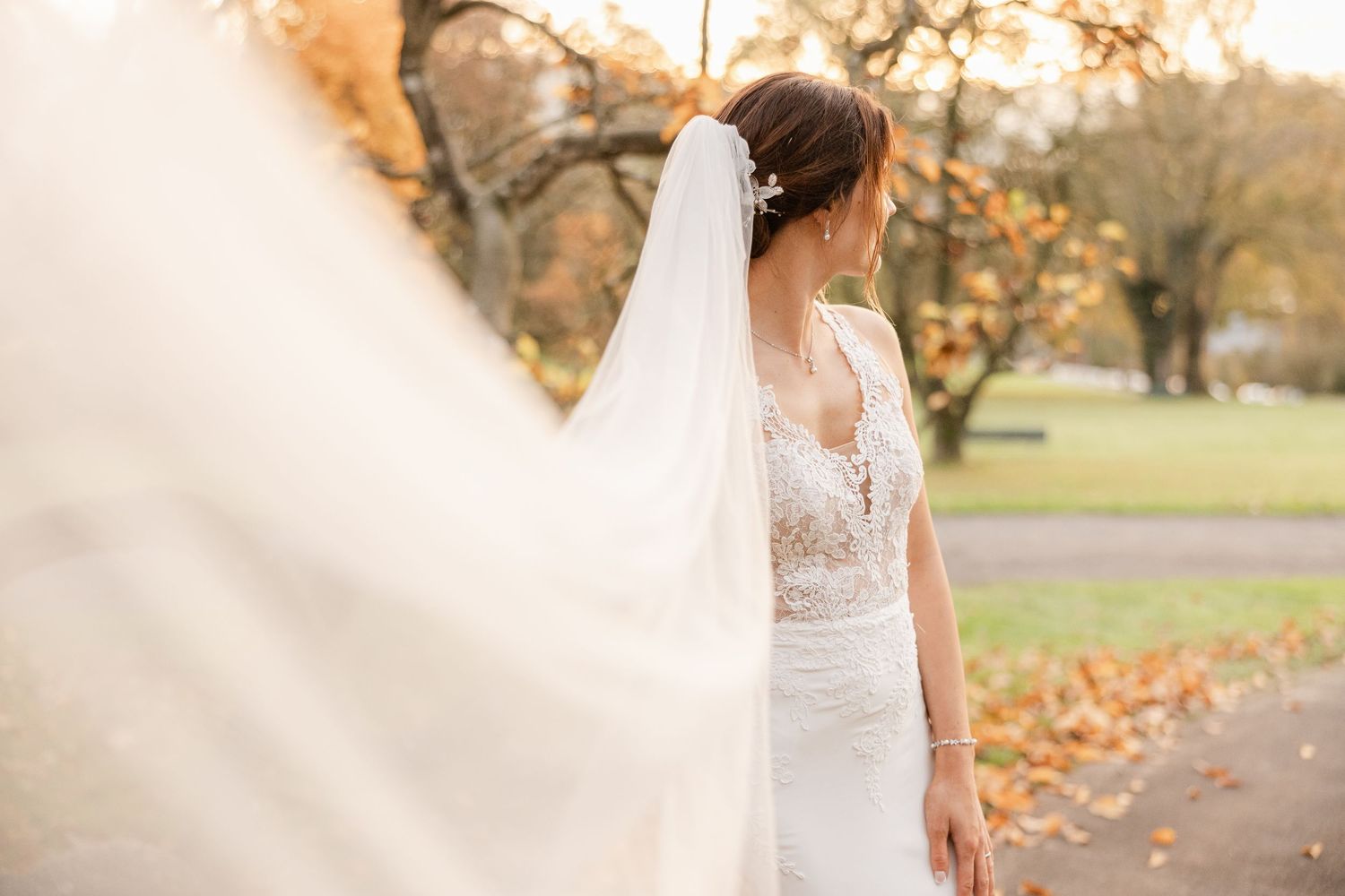 A bride in a lace wedding dress and long veil stands in an autumn park with golden leaves on the ground.