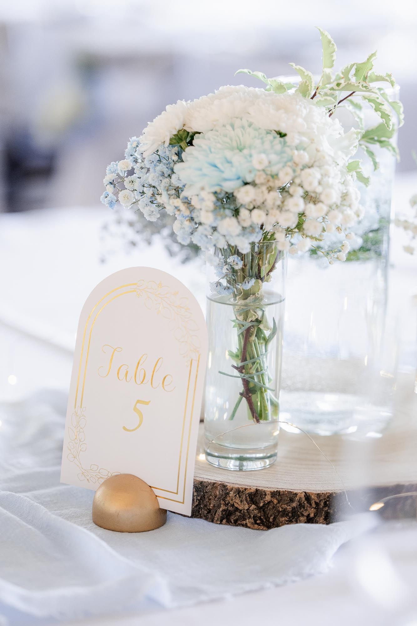 Wedding table 5 sign with gold lettering beside a glass vase of white and blue flowers on a wood slice centerpiece.