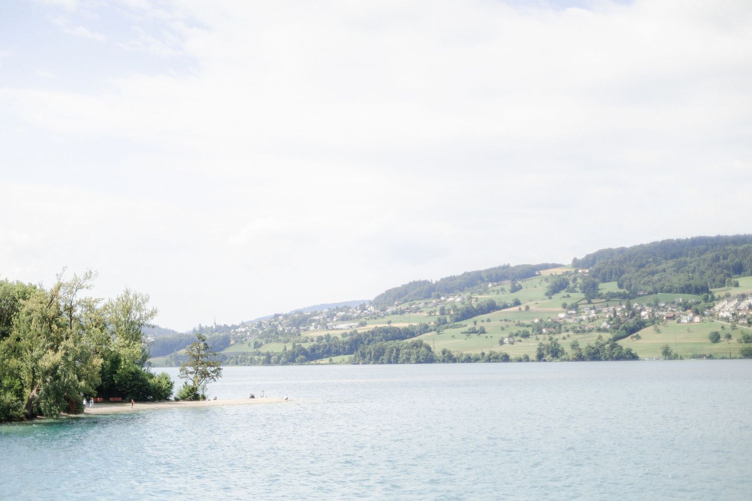 A panoramic view of a lake surrounded by green mountains and hills with sunny skies above.