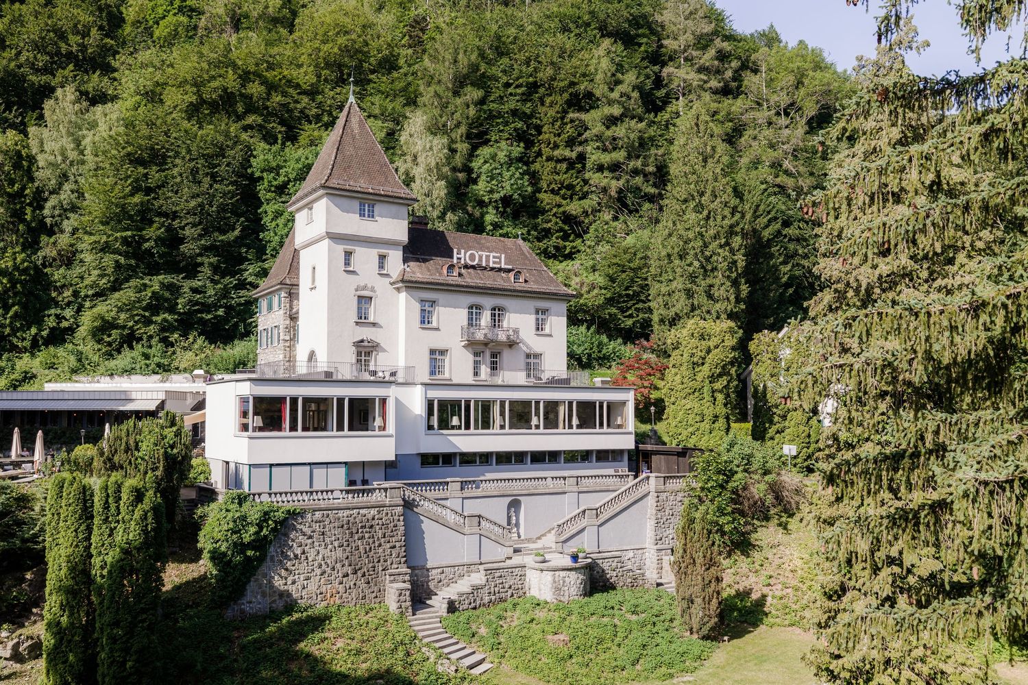 A historic white hotel building surrounded by lush green trees on a hillside, featuring a pointed tower and modern terrace extension.