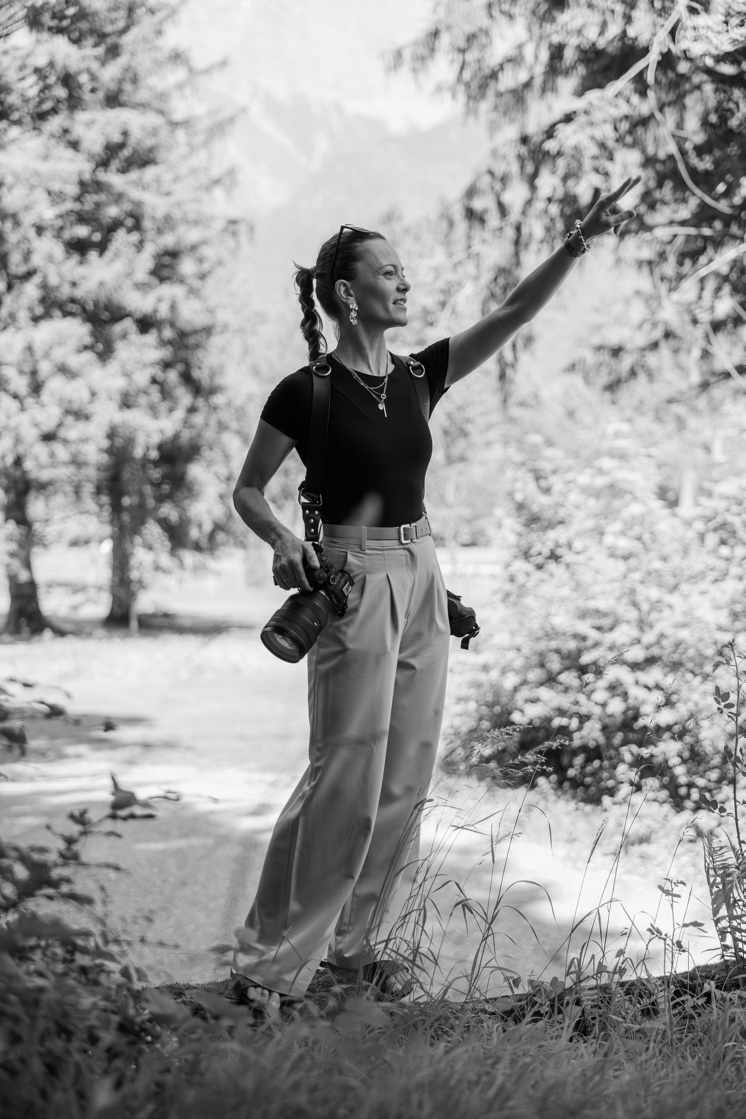Black and white photo of a woman holding a camera, pointing upward, standing on a forest path with mountains in the background.